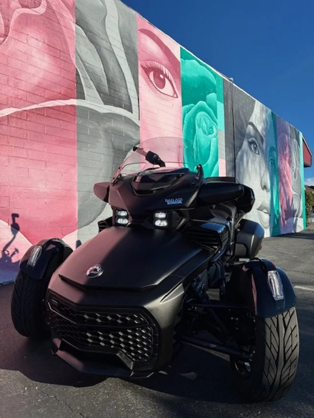 Black Can-Am Spyder three-wheeled motorcycle parked in front of a colorful mural featuring large, stylized portraits of women's faces and eyes.