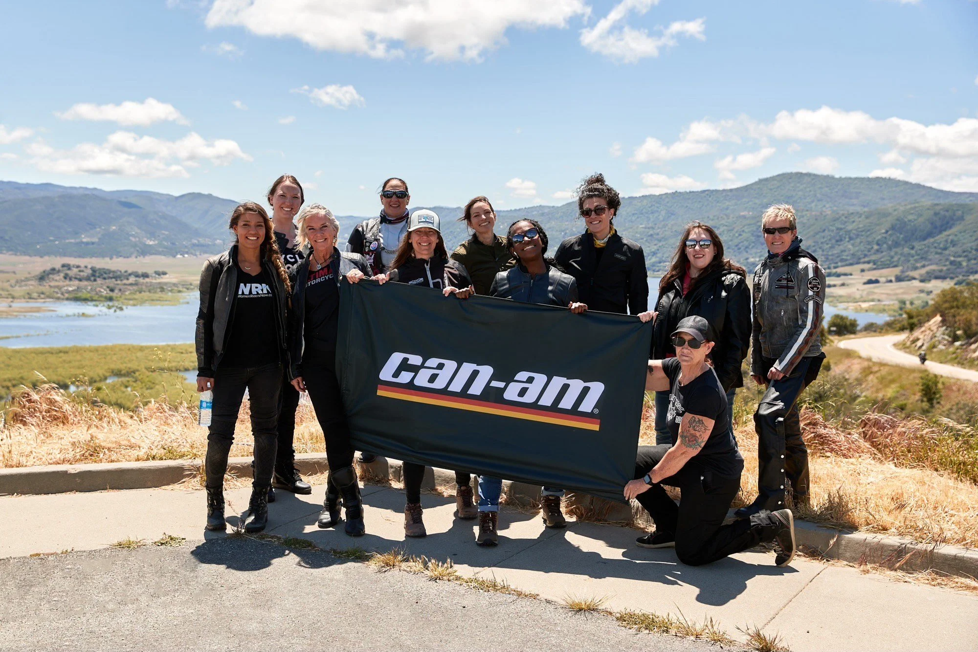 Group of women holding a Can-Am flag outdoors with mountains and water in the background on a clear day.
