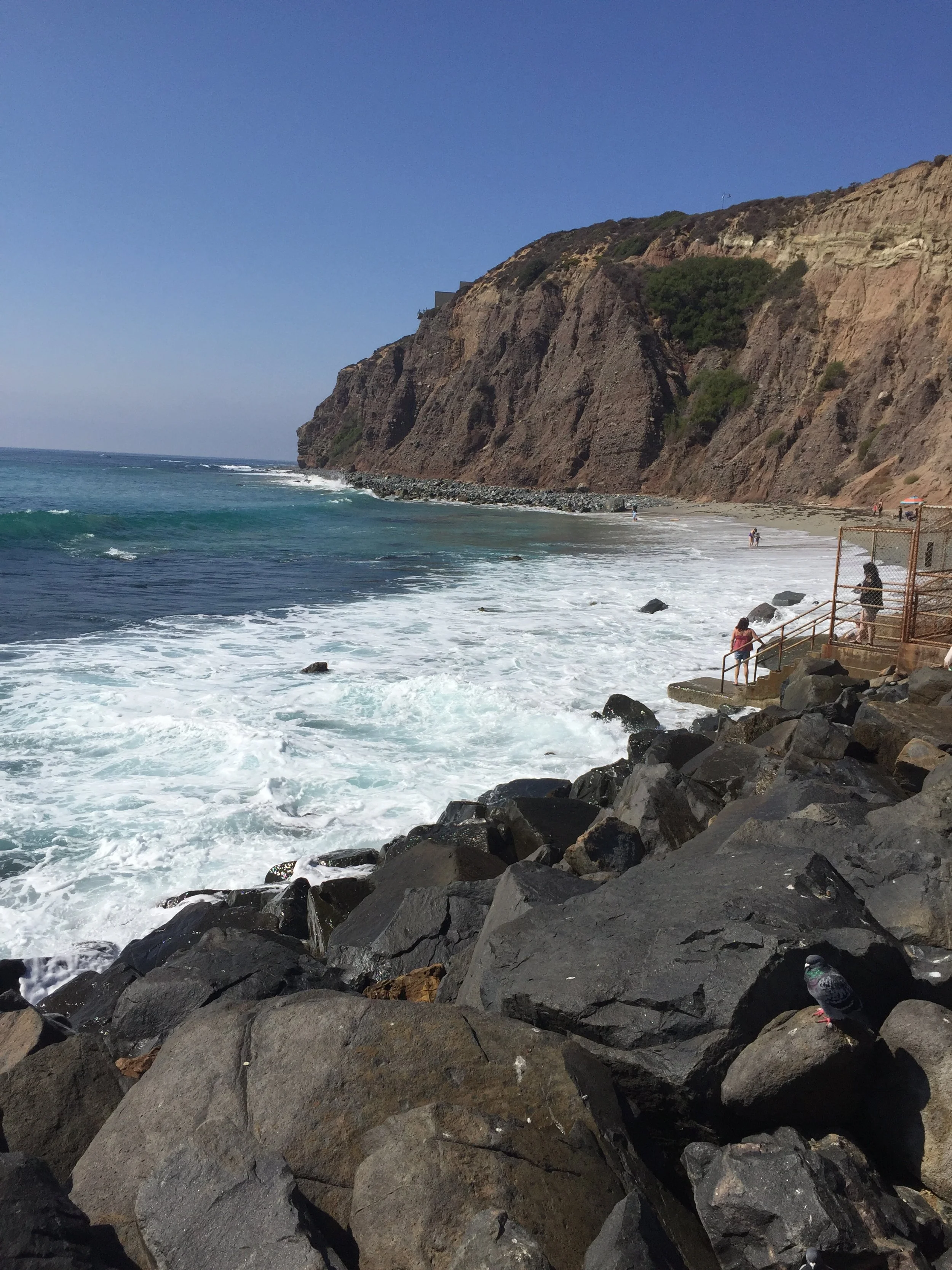 A rocky shoreline with waves crashing and a cliffside in the background, with a few people walking on the beach and stairs leading down to the water.