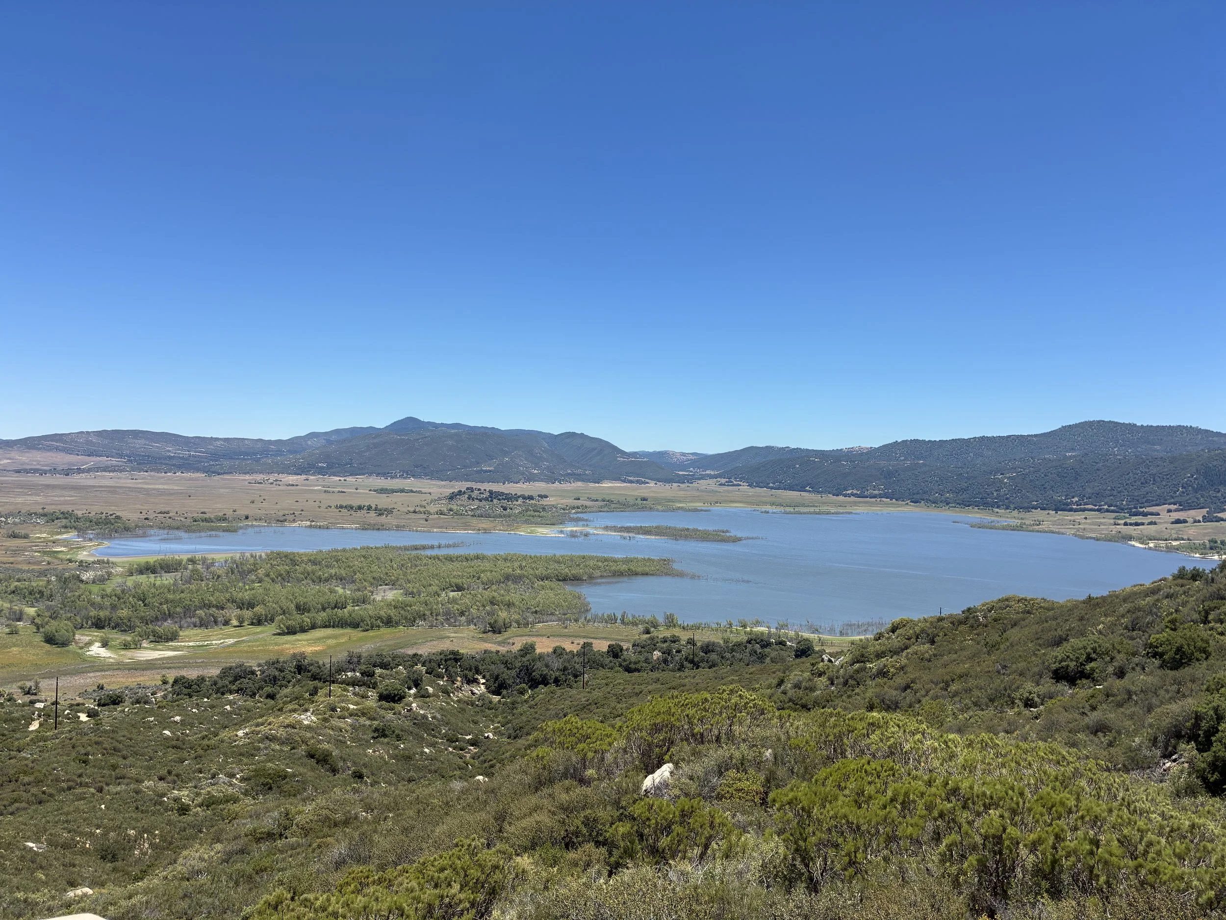 A landscape view of a lake surrounded by green hills and mountains under a clear blue sky.
