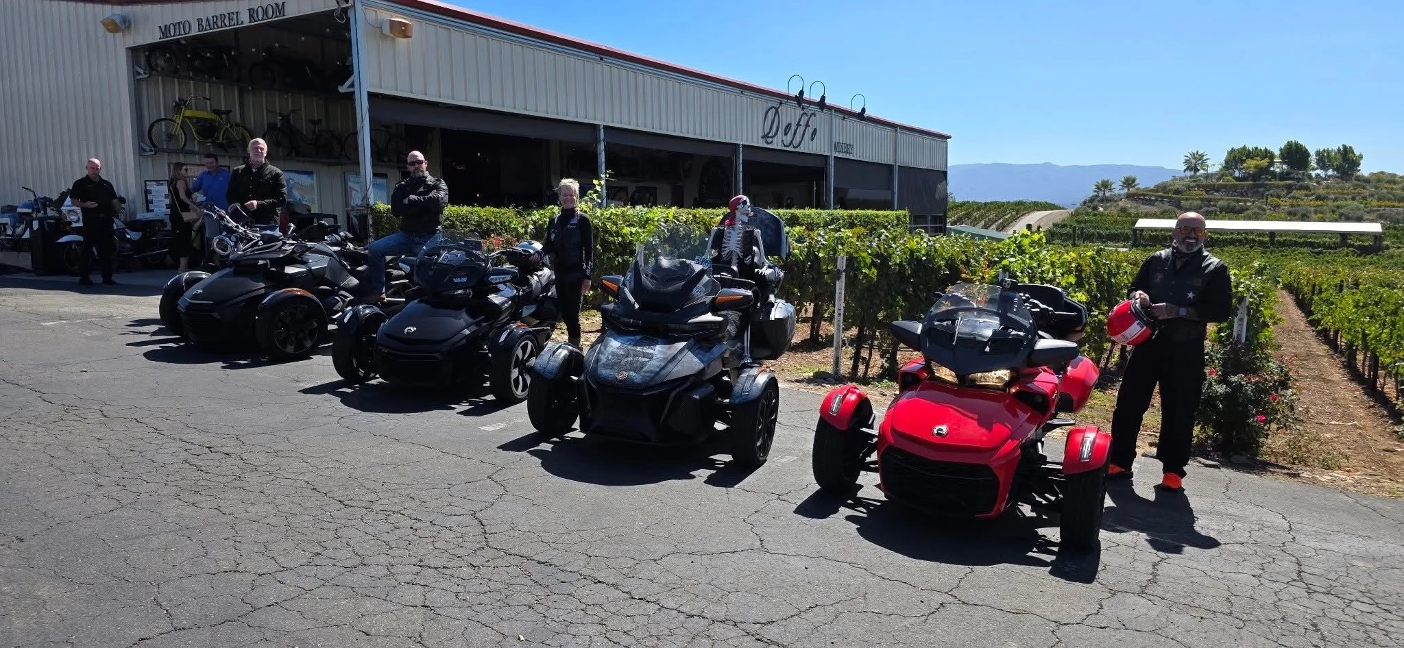 Group of people with motorcycles and trikes parked outside a building with a vineyard in the background.