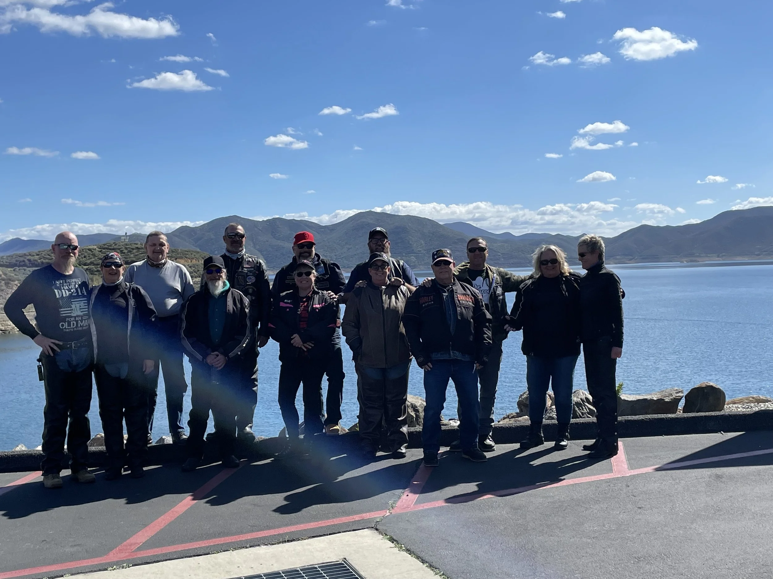 Group of people standing by a lake with mountains in the background, wearing casual and motorcycle clothing.