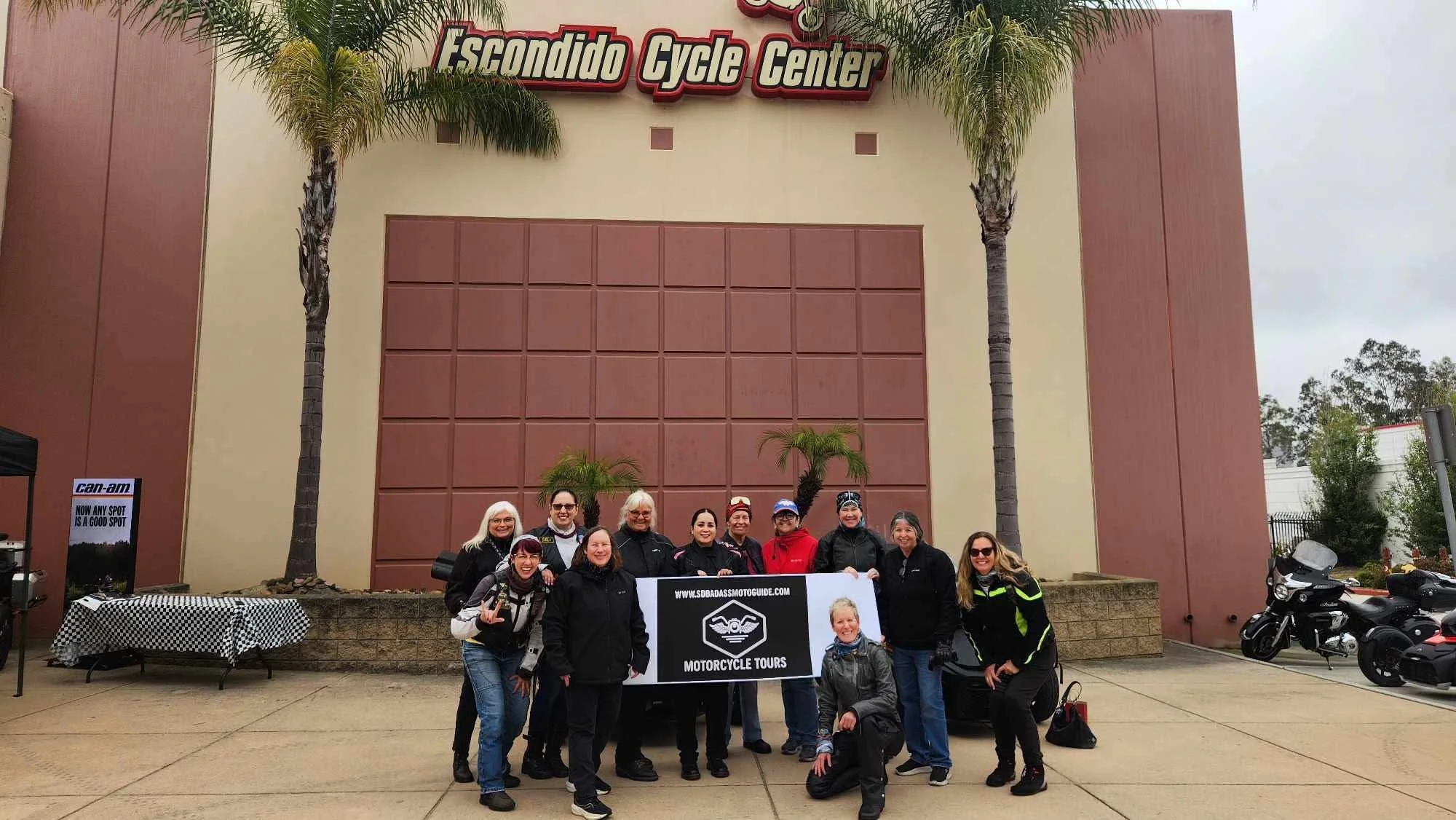 Group of people holding a motorcycle tour banner in front of Escondido Cycle Center, with palm trees and motorcycles in the background.