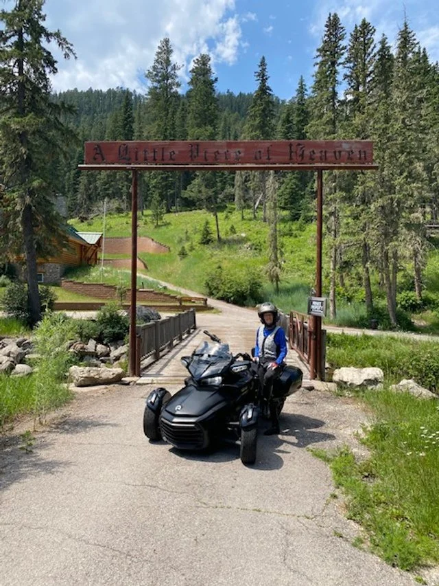 A person with a helmet sitting on a motorcycle in front of a bridge leading into a wooded area with a sign that reads, 'A Little Peer of Heaven'.