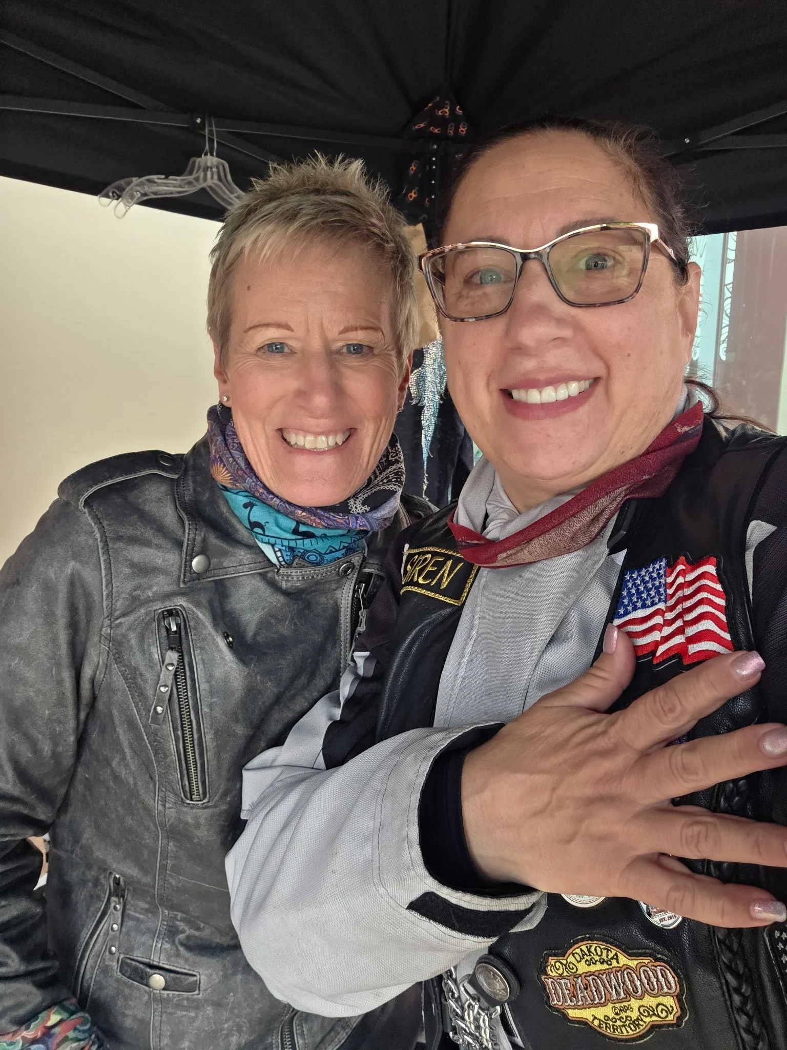 Two women smiling, one wearing a leather jacket and the other wearing a motorcycle vest with patches, one of which has an American flag patch, and they are taking a selfie together.
