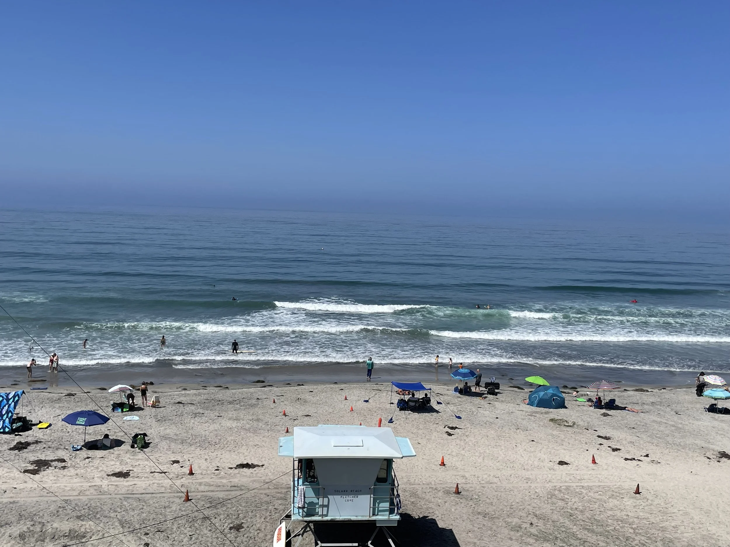 Beach scene with umbrellas, tents, and people in the sand and water under a clear blue sky.