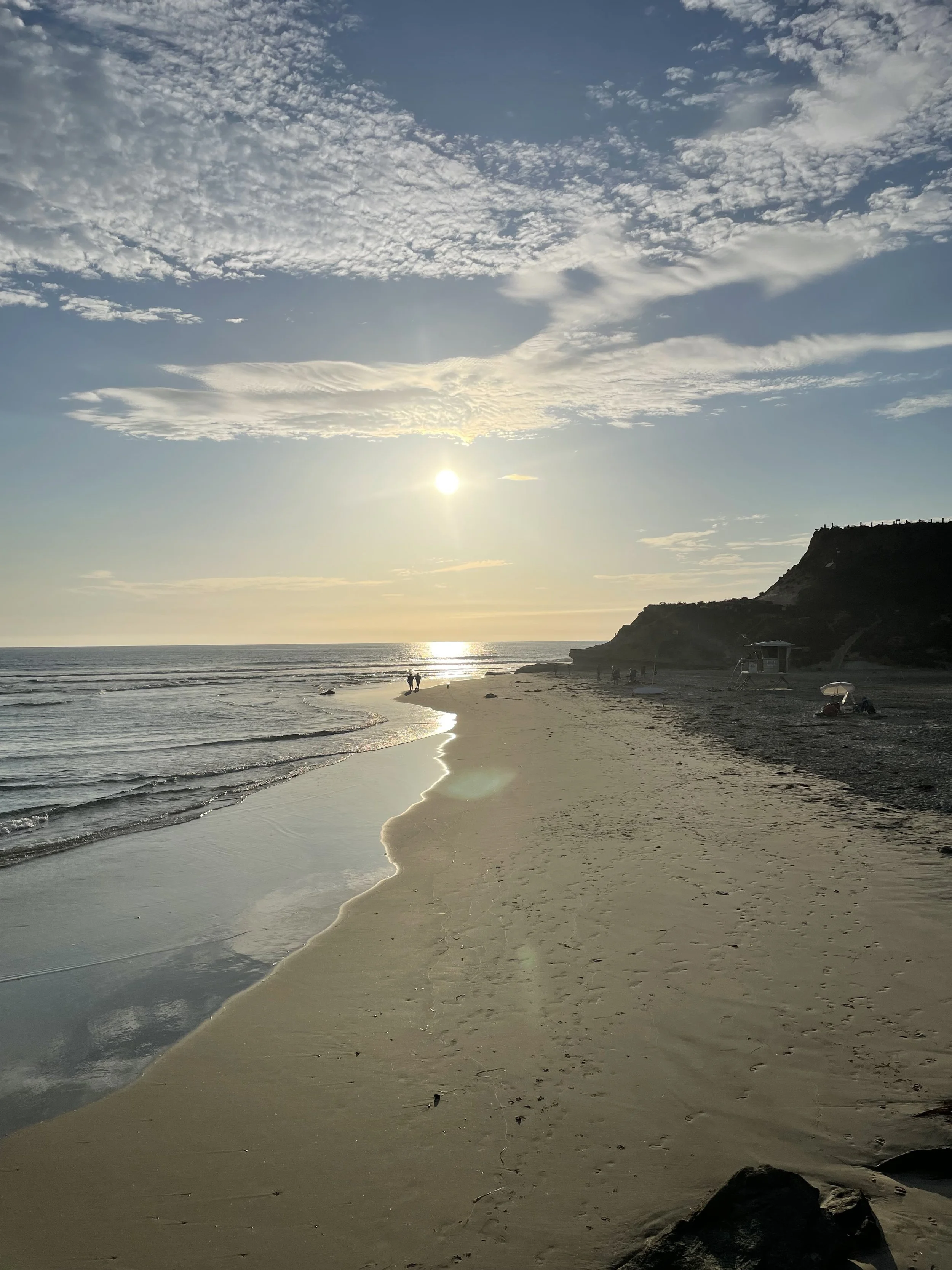 Sunset at a beach with a few people walking along the shoreline, rocky cliffs on the right, and a partly cloudy sky.