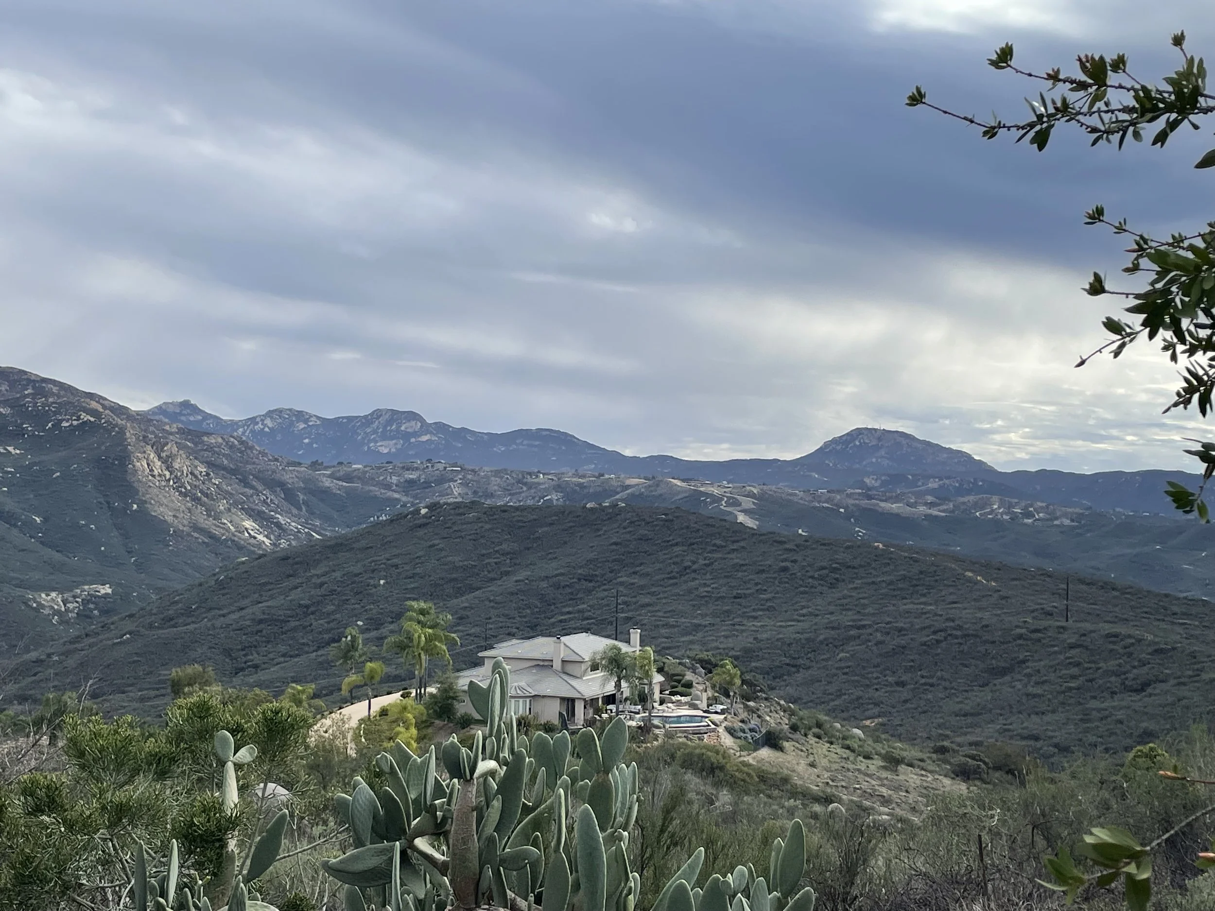 Scenic view of a mountainous landscape with a house and swimming pool on a hillside, desert plants in the foreground, under a cloudy sky.
