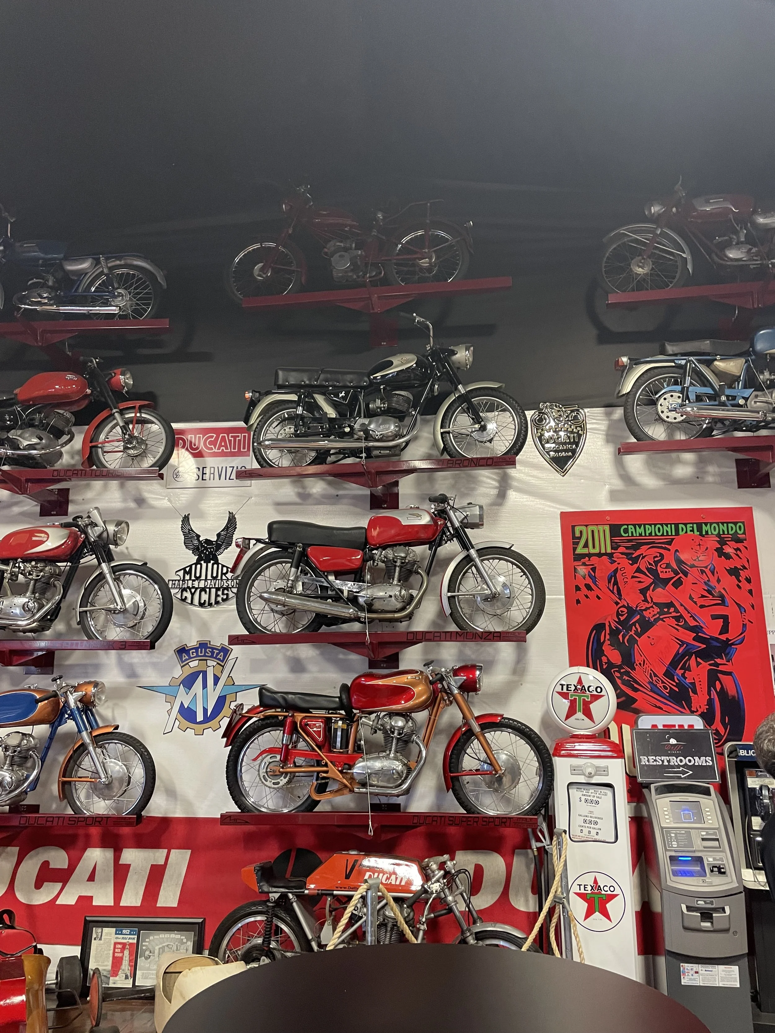 A display of vintage motorcycles mounted on a wall at a museum or collection, with various brands including Ducati, Harley-Davidson, and other classic models, along with vintage posters and signs.