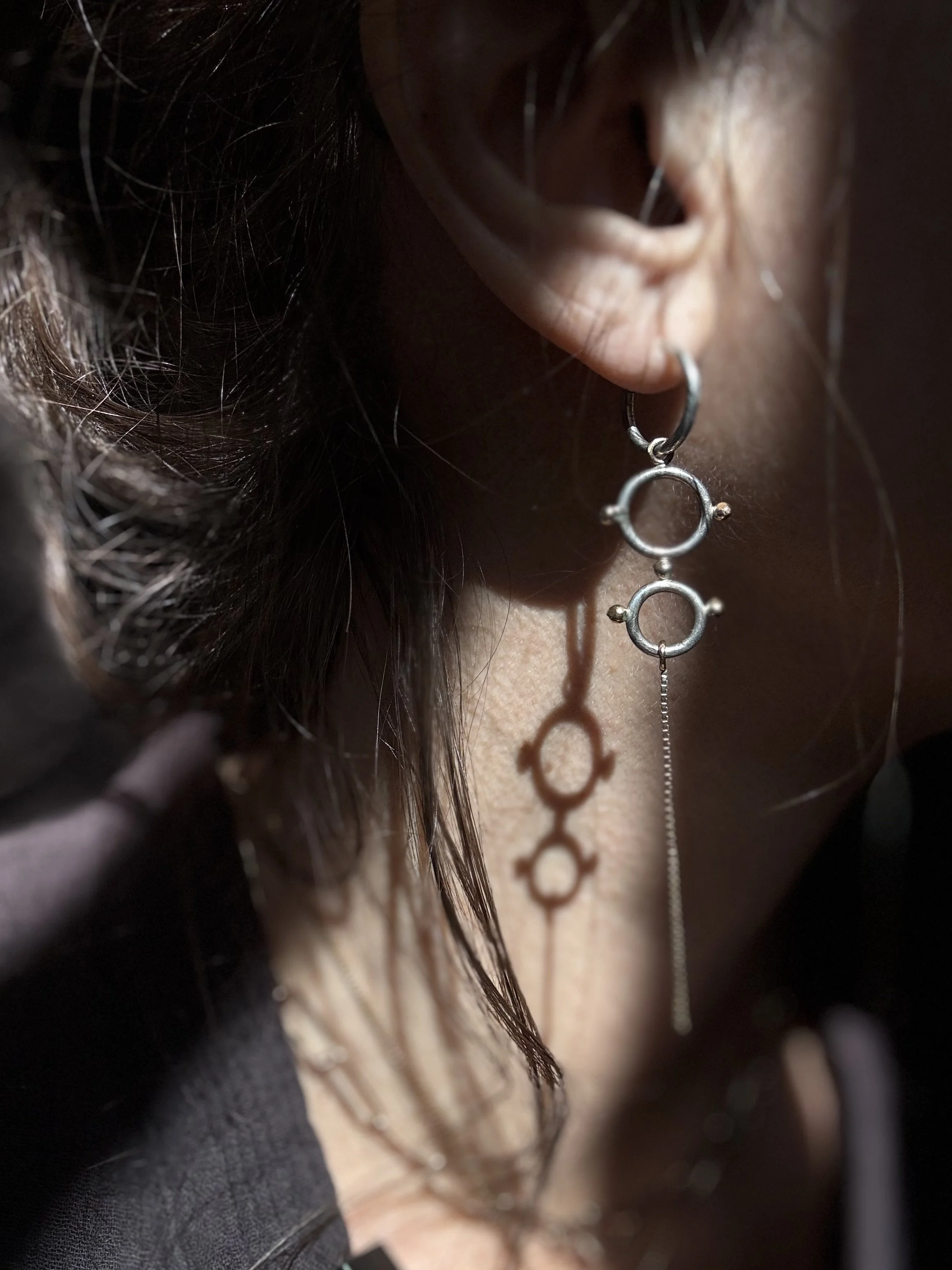 Woman wearing long silver earring featuring interconnected circles and a chain dangle, moody, classic and elegant casting a shadow on the neck.