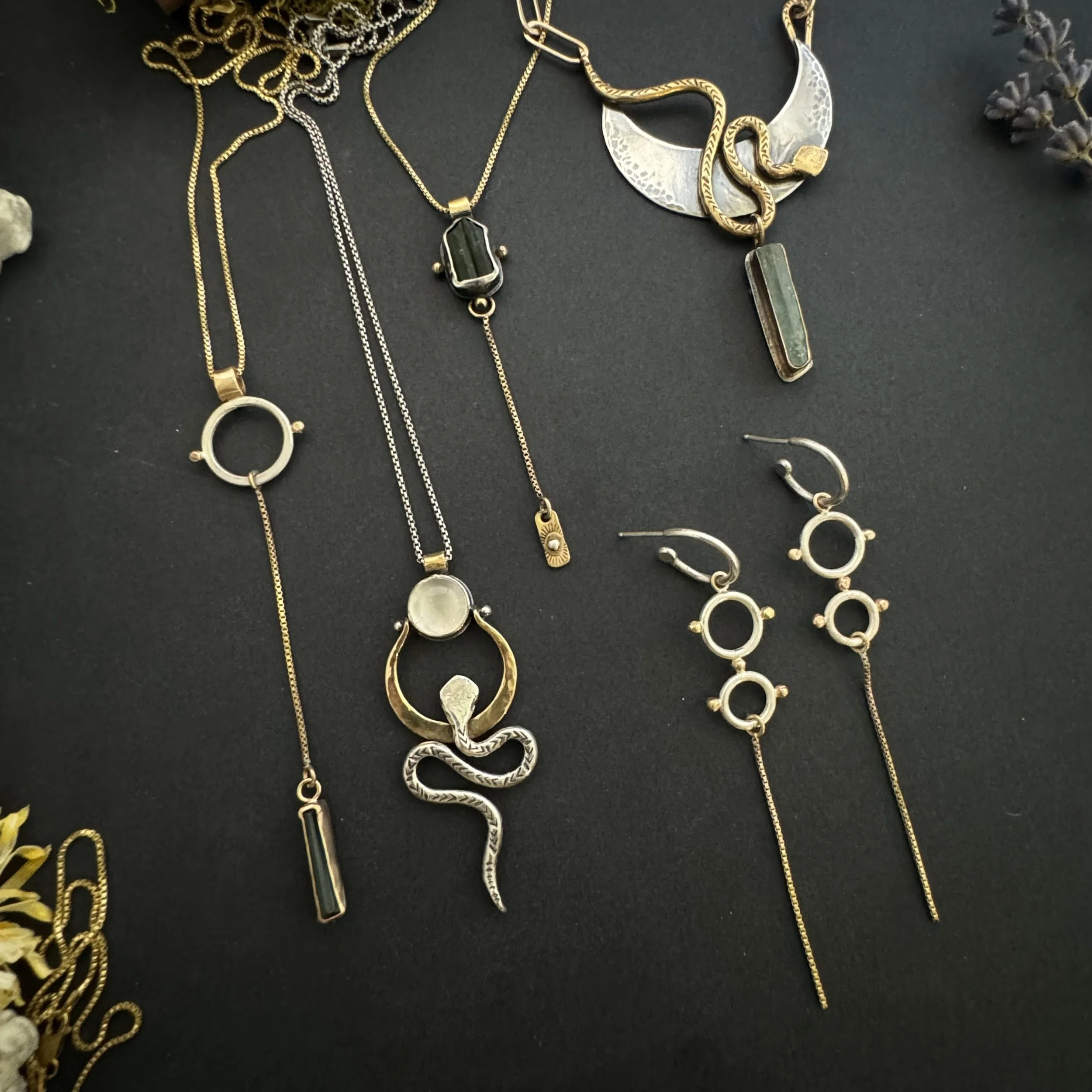 Various gold and silver necklaces and earrings with geometric and snake designs are arranged on a black surface.