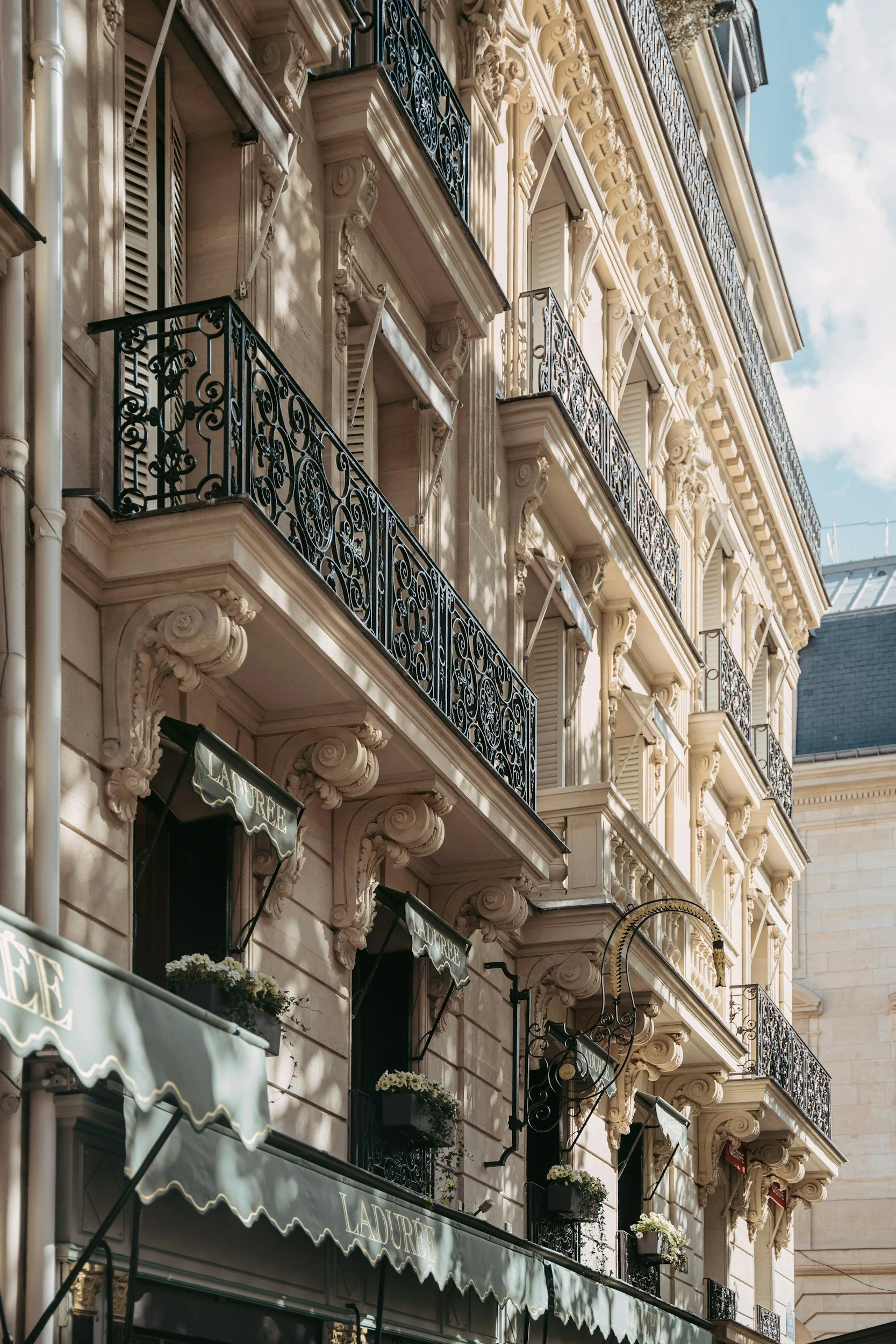 Hôtel avec architecture haussmannienne avec plusieurs balcons en fer forgé noir et fenêtres à volets blancs dans une rue ensoleillée.