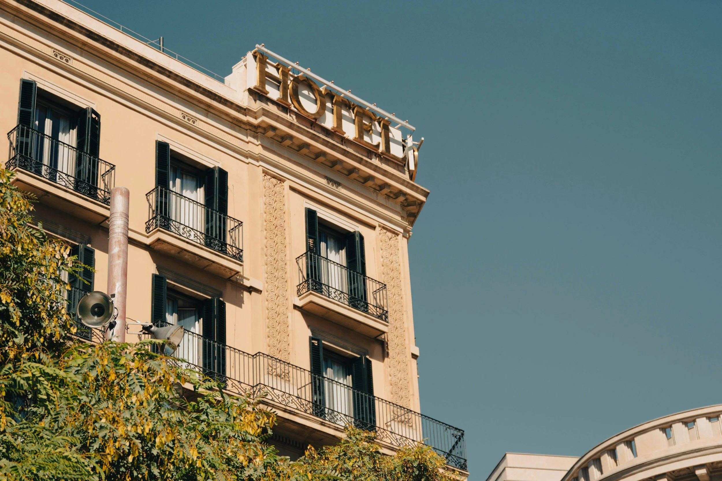 Facade d'un hôtel en France avec un grand signe 'HOTEL' au sommet, fenêtres à volets noirs, balcons en fer forgé, arbres en premier plan et ciel bleu en arrière-plan.