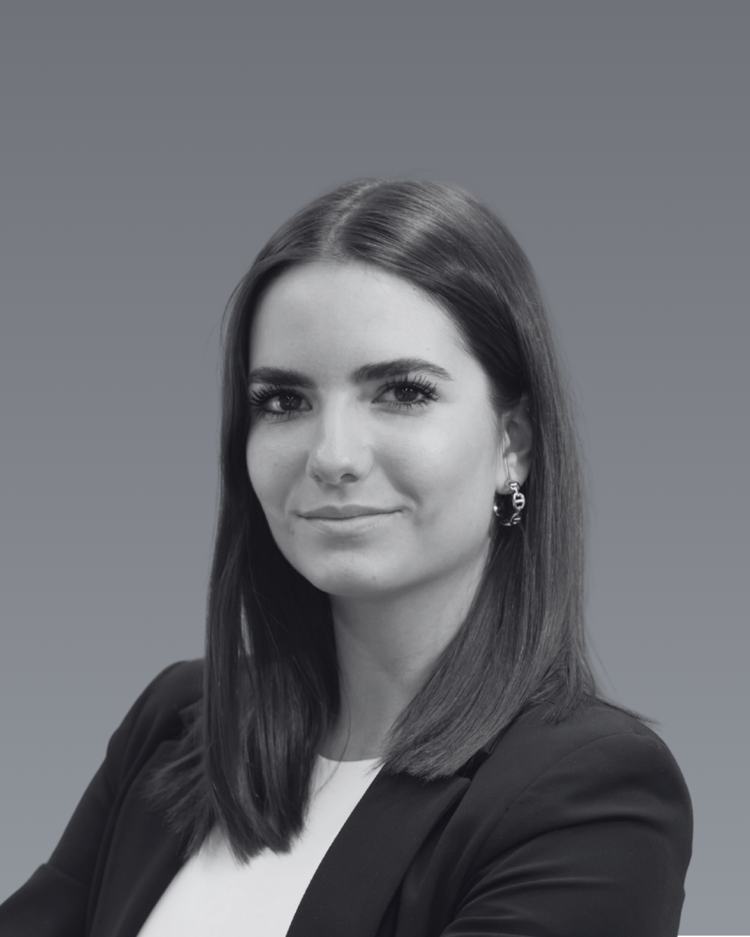 Portrait en noir et blanc d'Alicia Loubat aux cheveux bruns, portant un blazer noir, avec un fond gris unicolore.