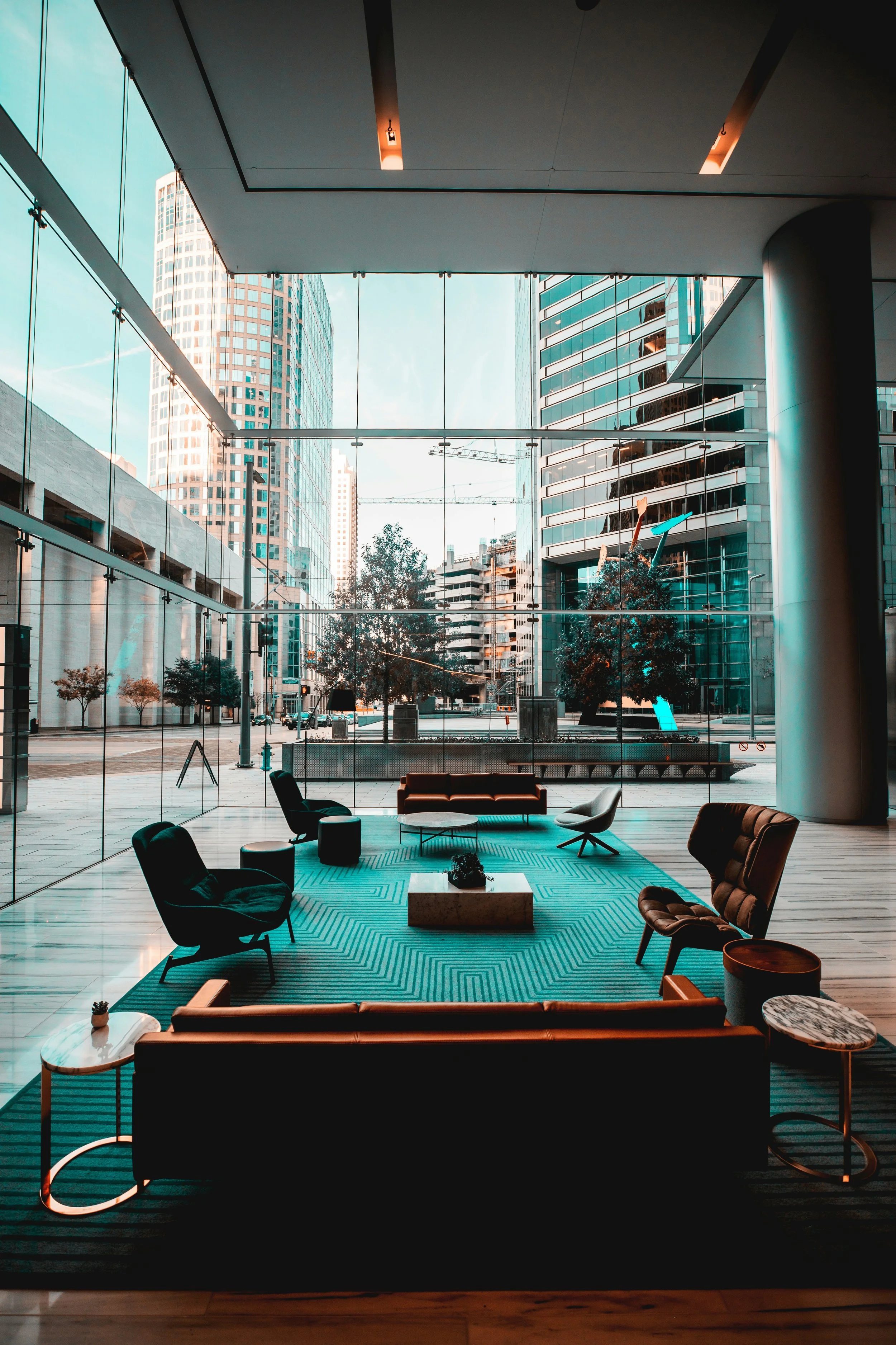 Hall d'hotel moderne avec des fauteuils et canapés noirs autour d'un tapis turquoise dans une salle de réception ou salle de réunion, avec de grandes fenêtres offrant une vue sur des bâtiments urbains modernes.