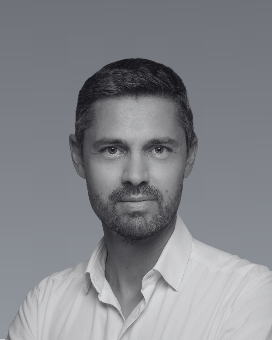 Portrait noir et blanc de Stéphane Lamblin avec une barbe, portant une chemise blanche, regardant directement l'objectif, fond gris