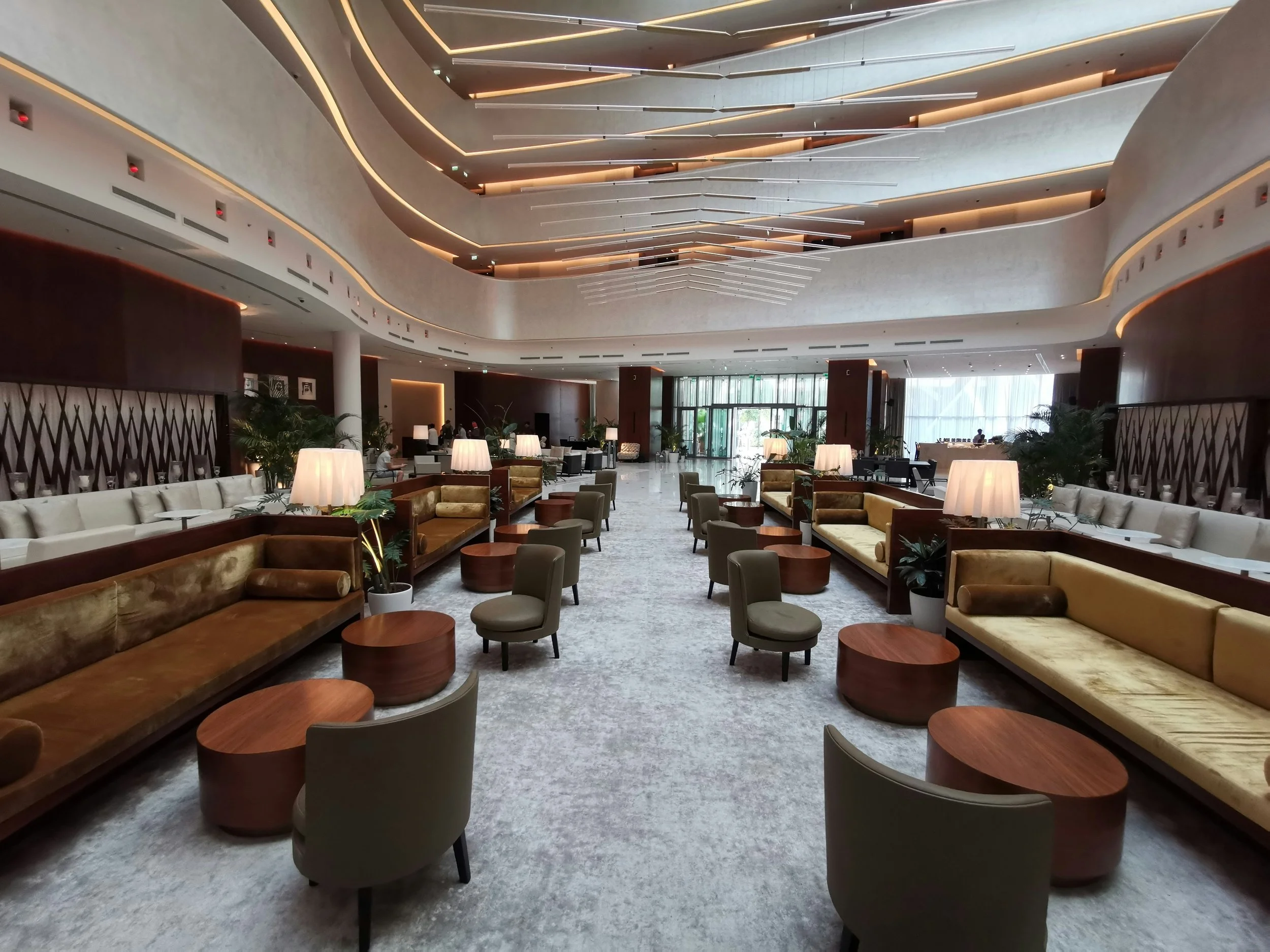 Un grand hallélégamment décoré avec des canapés en velours beige et marron, des tables basses en bois, des lampes de table avec abat-jour blanc, des plantes en pot, et de grands vitres permettant la lumière naturelle. L’intérieur présente un design moderne avec des étages ouverts et un mobilier confortable.