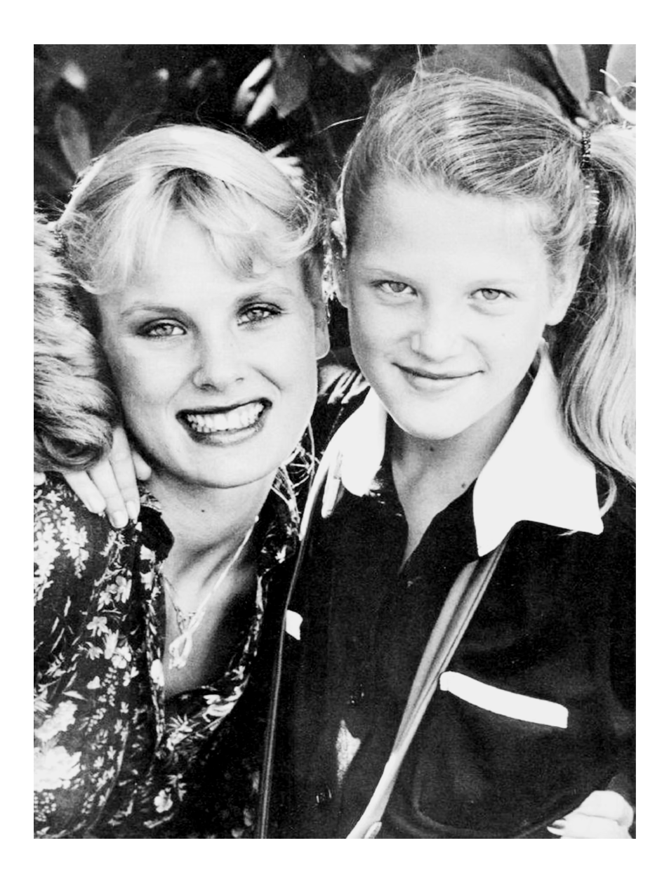 Black and white photo of two smiling women, one with curly hair and the other with straight hair tied back, standing close together outdoors.