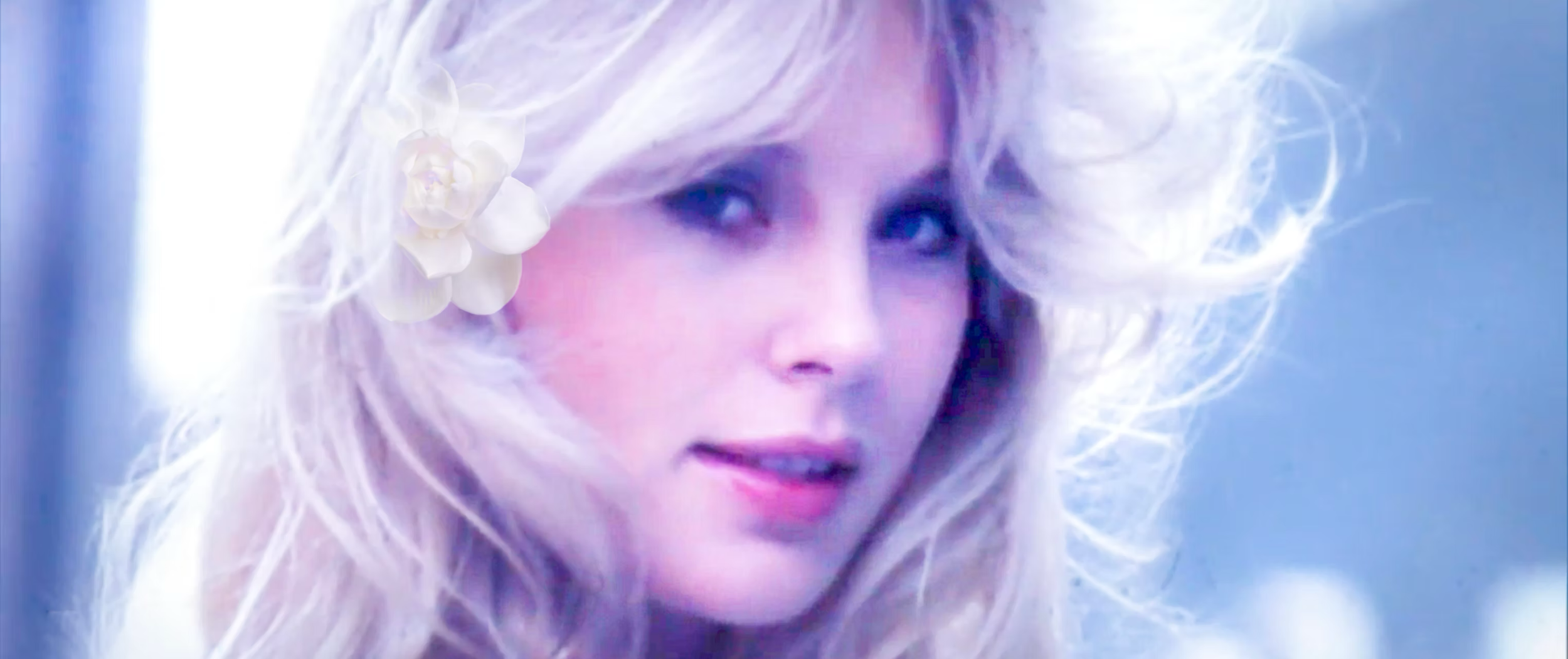 A woman with long, wavy blonde hair adorned with a white flower, looking slightly to the side with a soft expression, in a bright, pastel-lit setting.