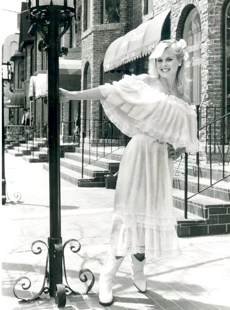 A woman with blonde hair, wearing a vintage-style dress and white boots, smiling and holding an umbrella, standing on a city sidewalk with brick buildings in the background.
