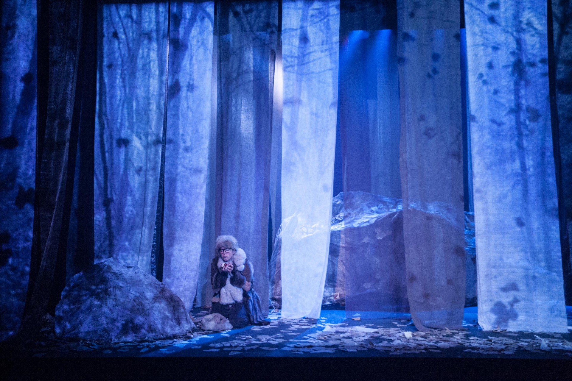 A person in costume sitting on the ground in a theatrical scene with blue lighting, surrounded by trees and large rocks, holding something in their hands.