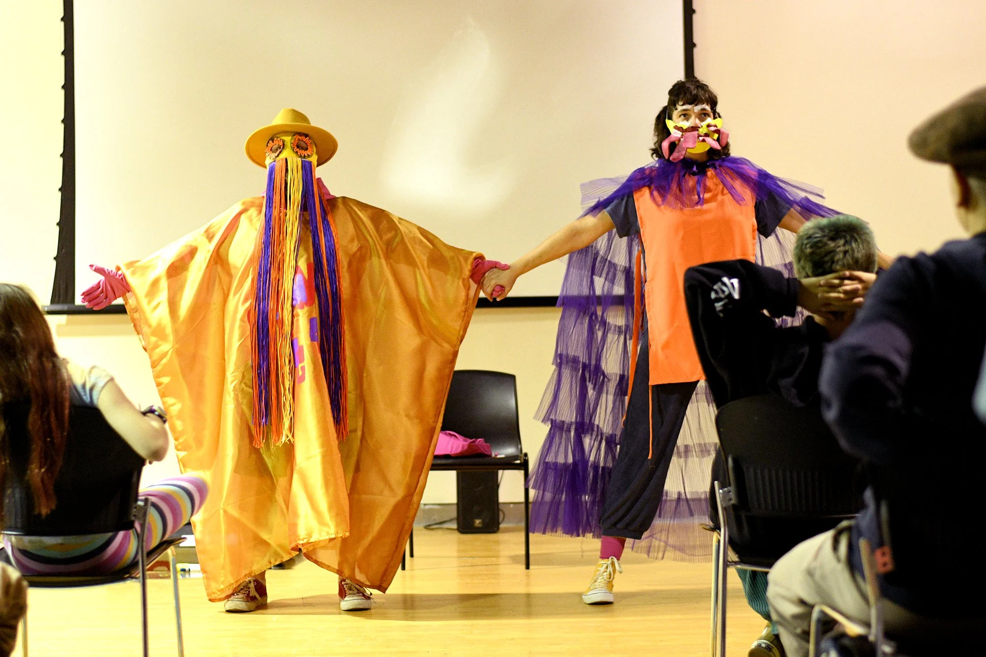Two performers dressed in colorful costumes on a stage, holding hands, with an audience seated in front. One performer wears an orange and purple outfit with a mask, and the other wears a large yellow cape, a wide-brimmed hat, and a mask with long, multicolored strands.