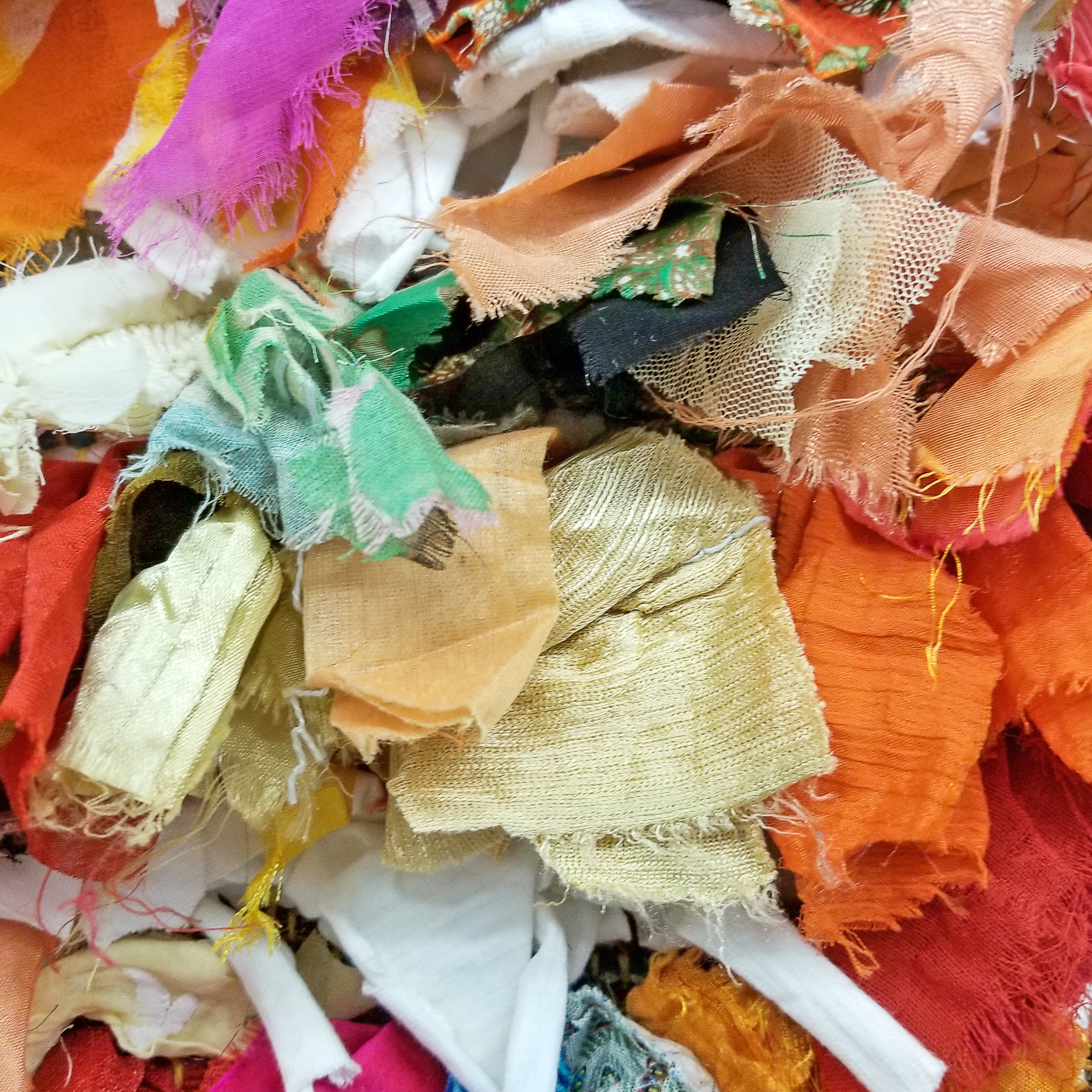 A close-up view of a tangled pile of colorful fabric scraps, including various textured and patterned pieces in shades of orange, pink, green, gold, black, and white.
