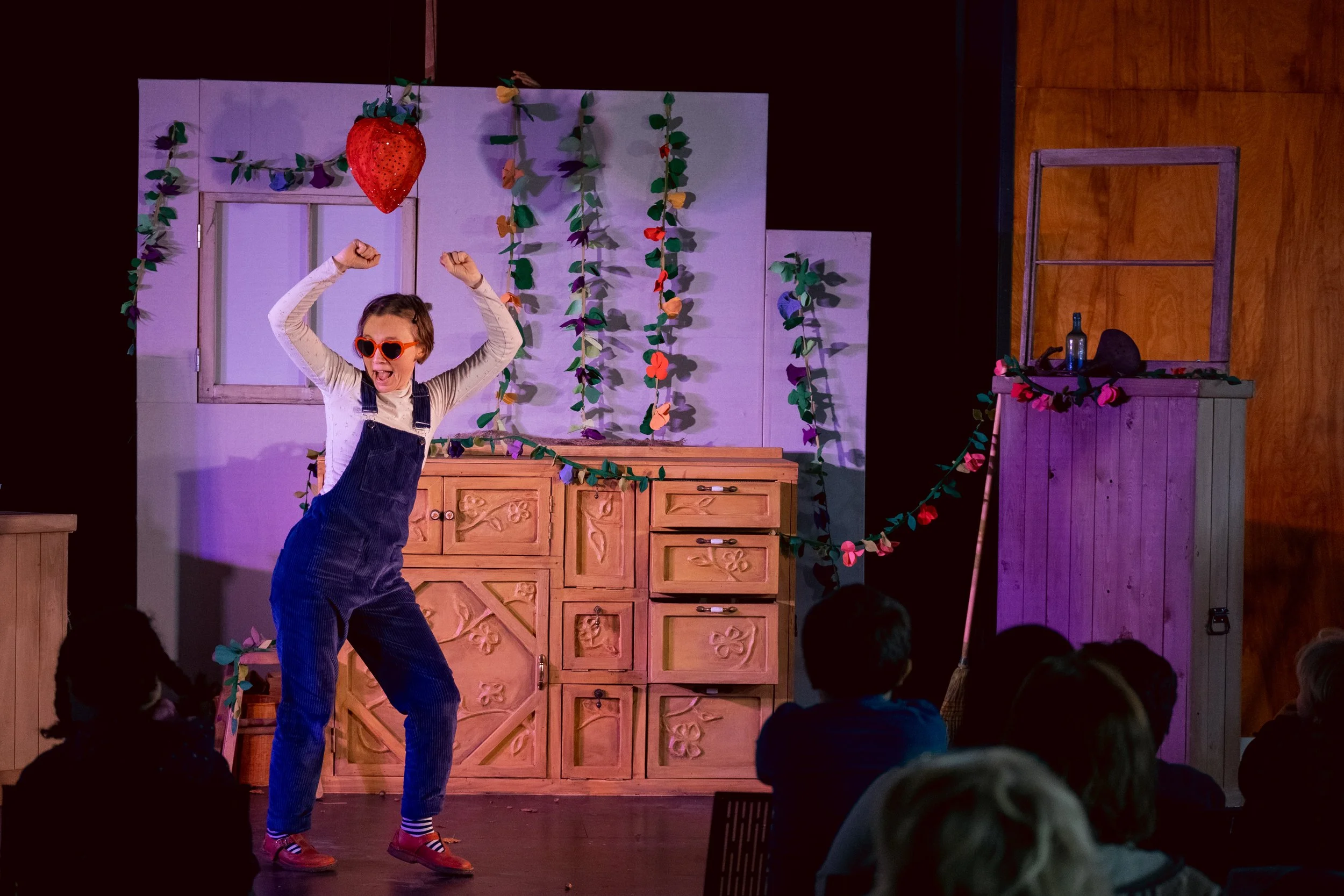 A young girl with glasses and overalls excitedly performing on stage in front of an audience, with colorful decorations and a wooden backdrop.