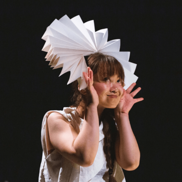 Young girl with long brown hair and bangs holding her hands near her face, with a playful expression. She has a large, white, origami paper hat on her head.