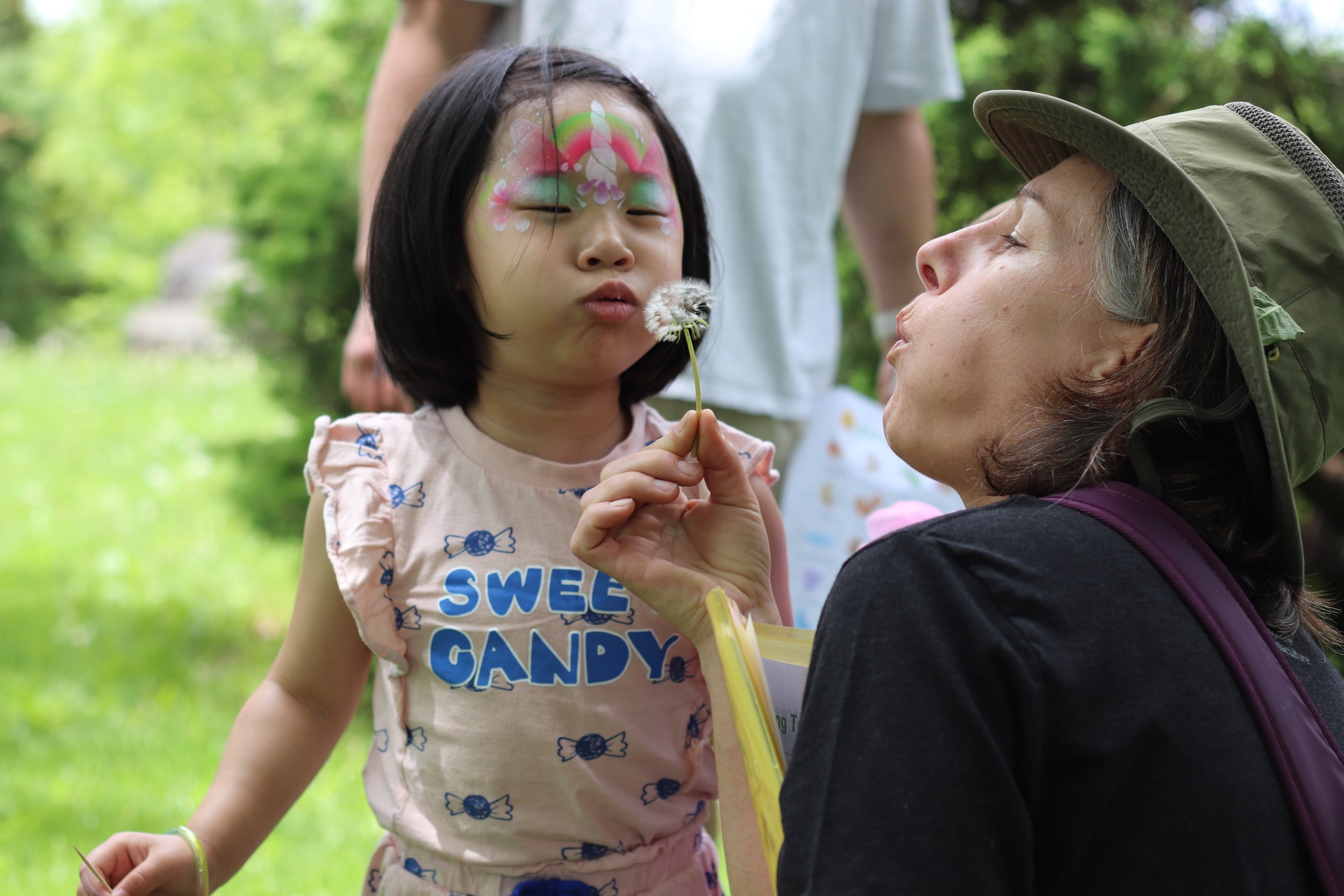 Young girl with face painted as a butterfly blowing dandelion with a woman wearing a hat