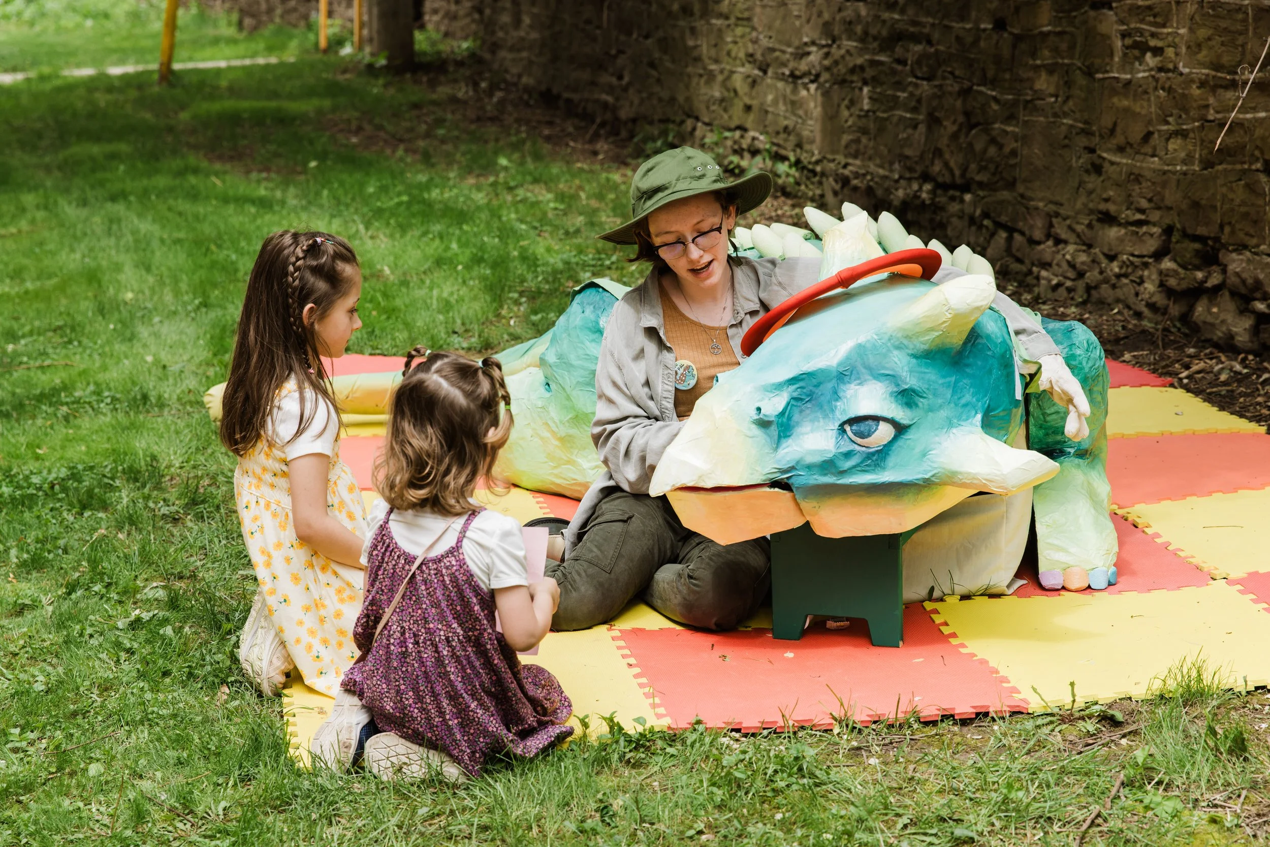 A woman with two young girls, sitting on a colorful foam mat outdoors, explaining a dragon puppet to them.