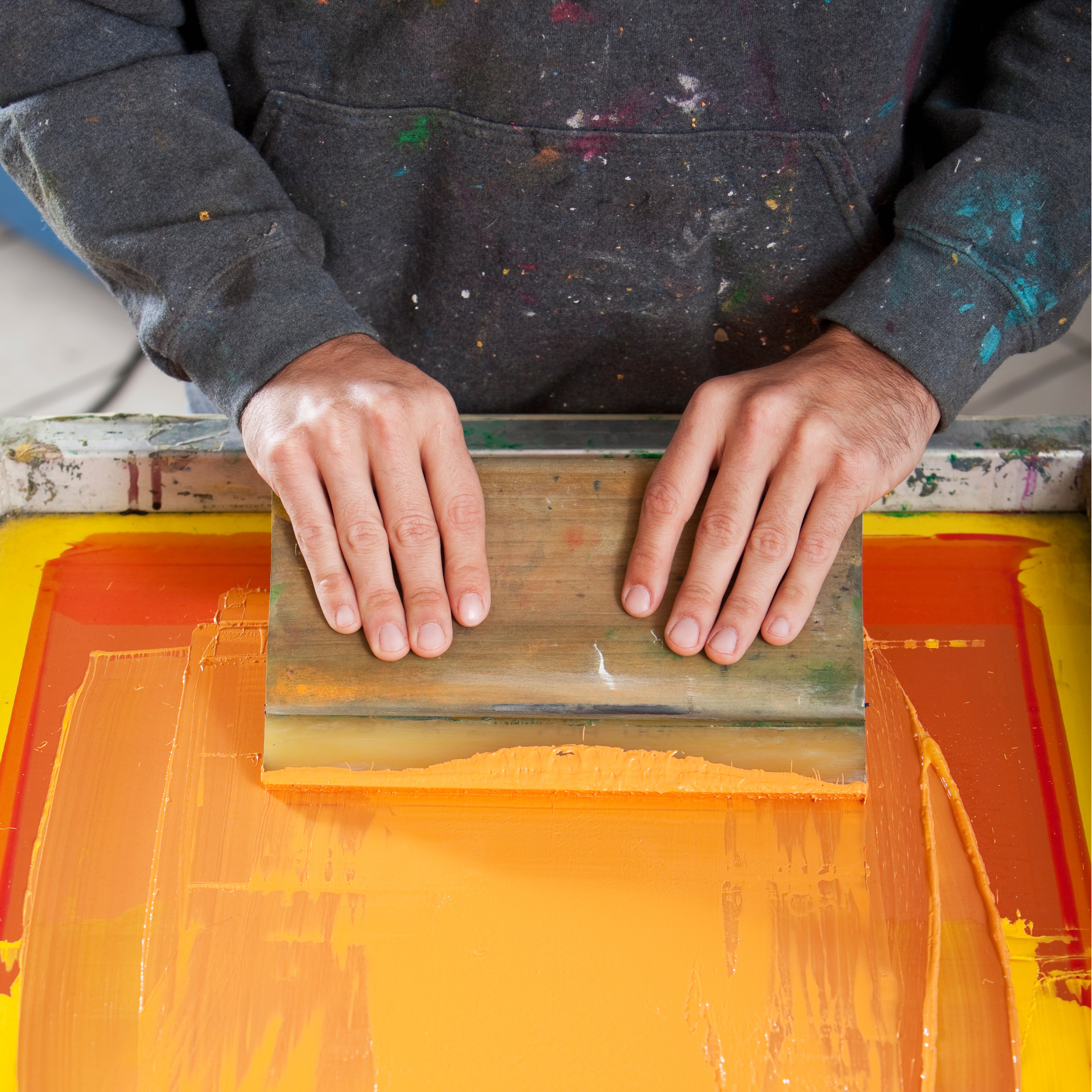 Person in a black, paint-splattered hoodie using a squeegee to spread orange ink on a screen for printing.