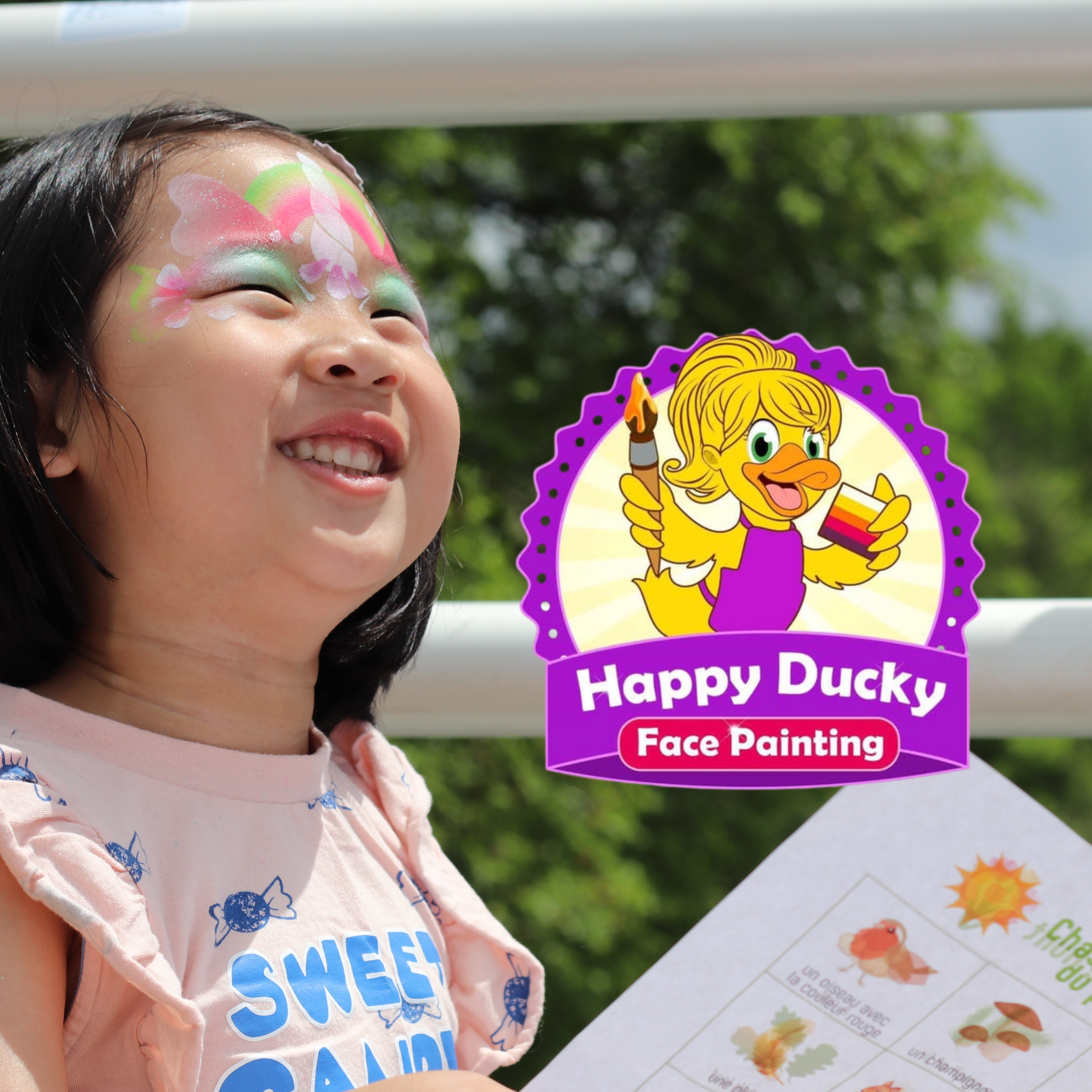 Smiling girl with face paint of pink and green flowers, at outdoor event with a 'Happy Ducky Face Painting' sign.