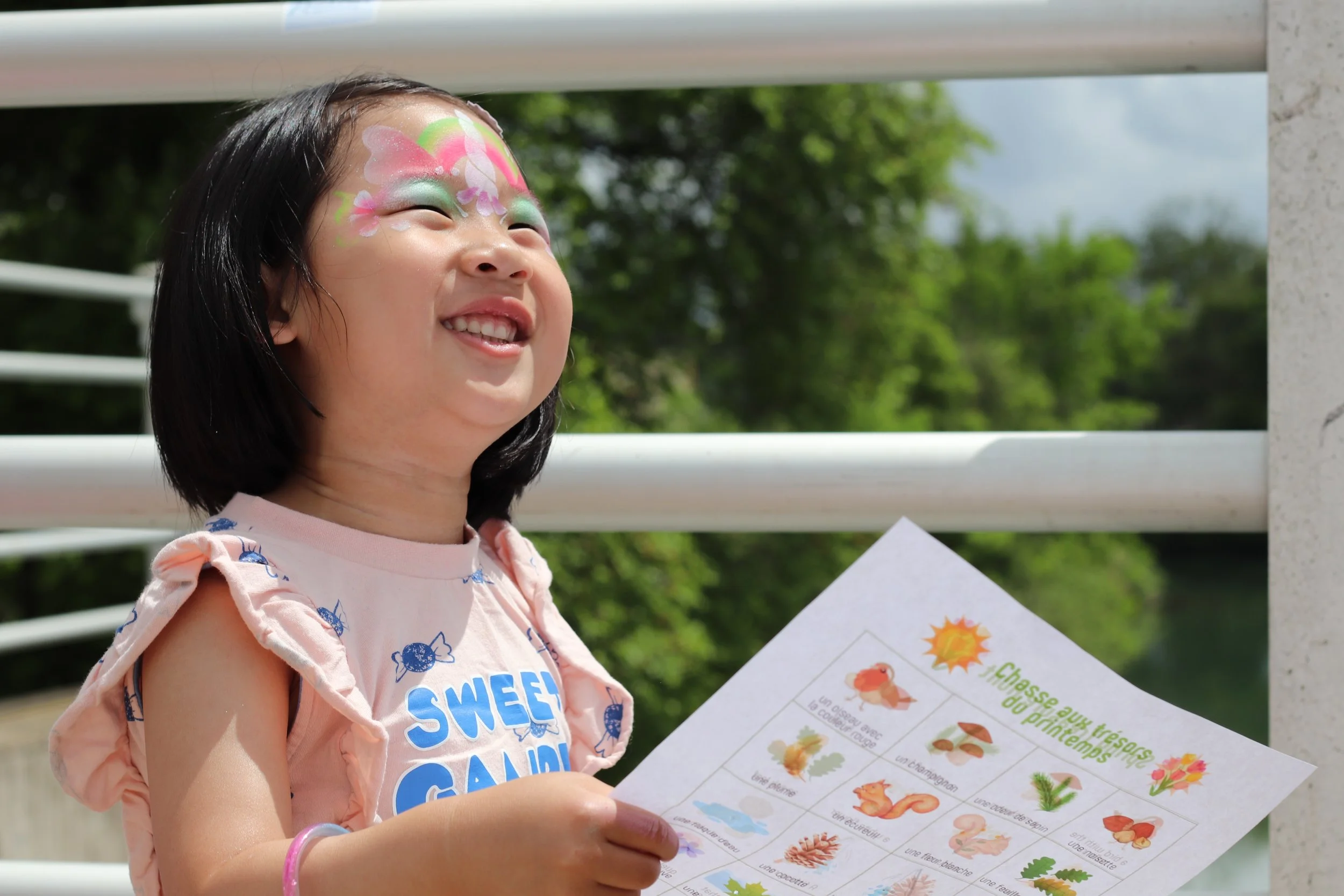 A young girl smiling outdoors, holding a colorful educational worksheet with painted floral face art and wearing a pink shirt.