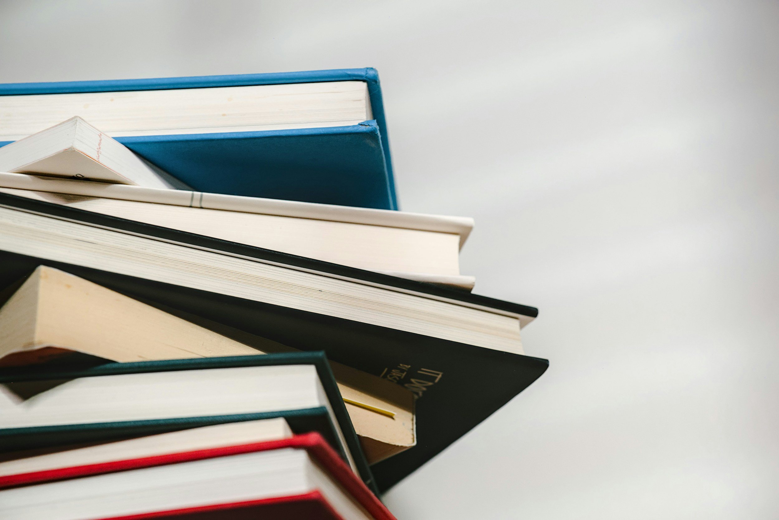 Close-up of stacked books with various colored covers on a white surface and a plain light background.