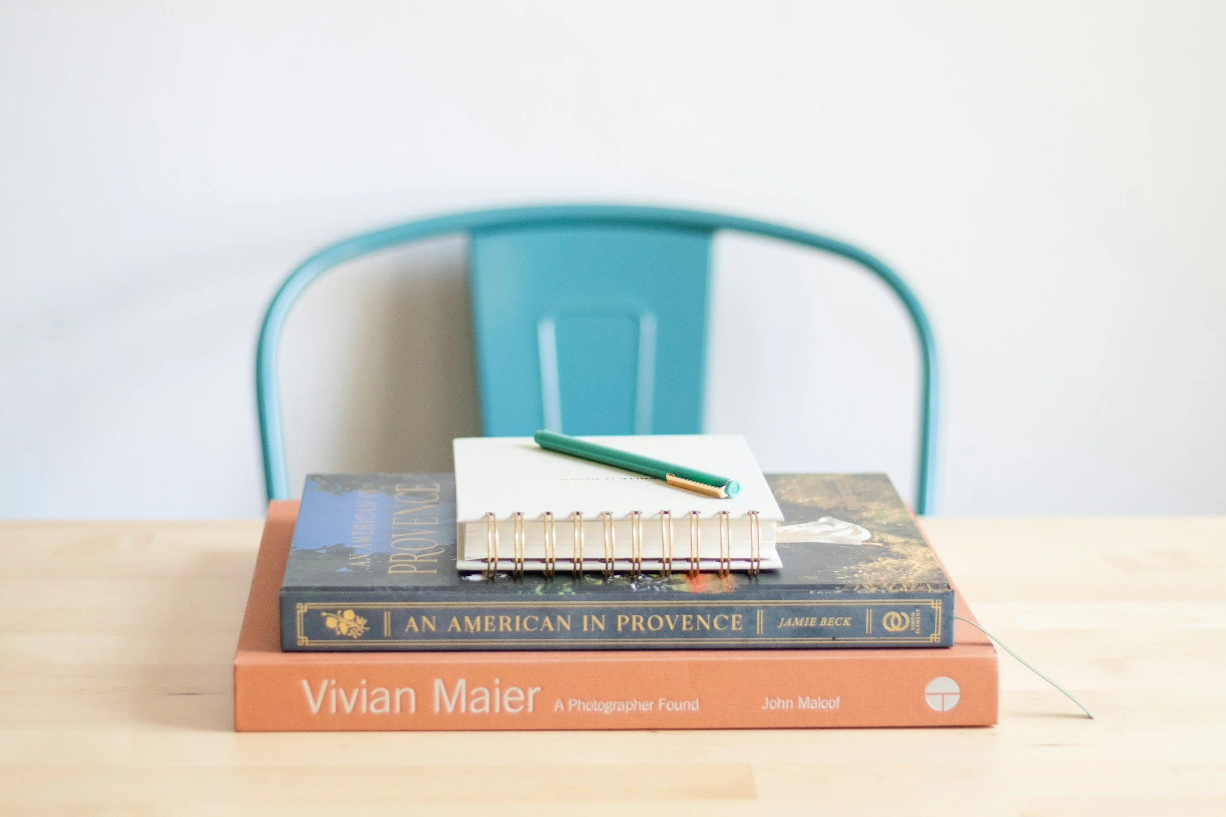 A stack of books on a wooden table with a teal chair in the background, and a notebook with a pen on top of the books.
