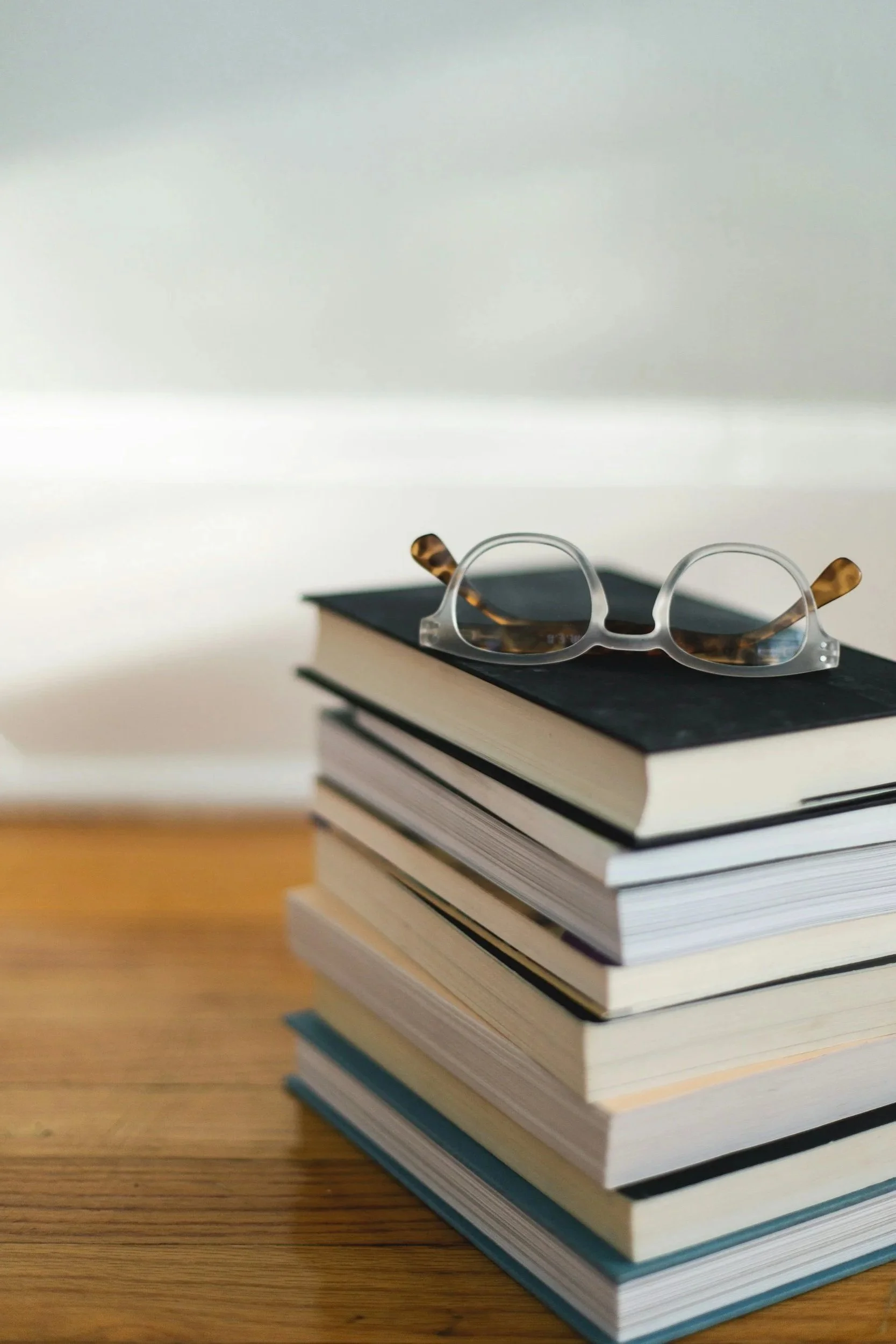 A stack of hardcover books and a pair of eyeglasses with tortoise-shell arms resting on top of a wooden surface.