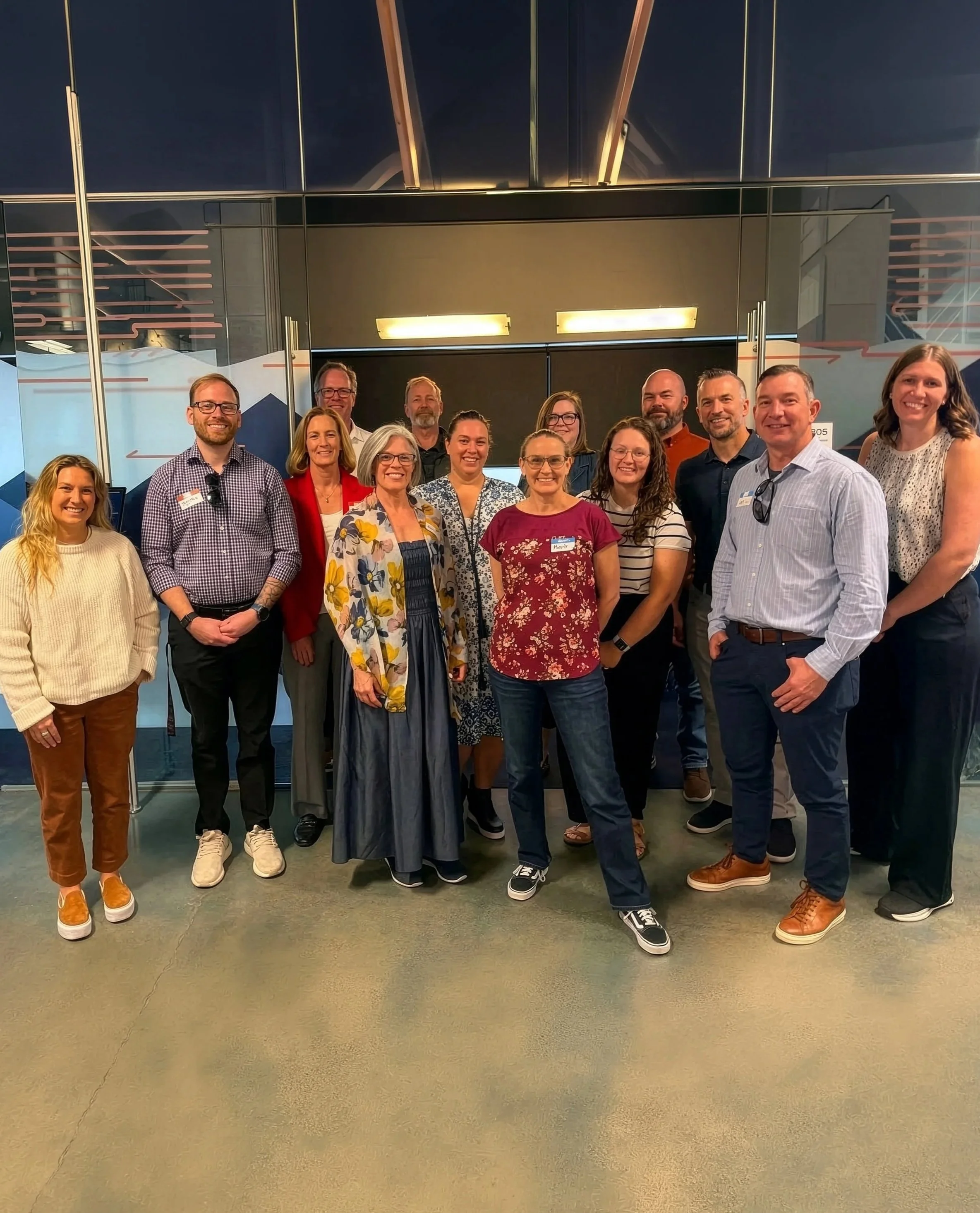 ENRG group of fifteen people posing together indoors, some wearing name tags, in front of glass walls with modern architectural features.