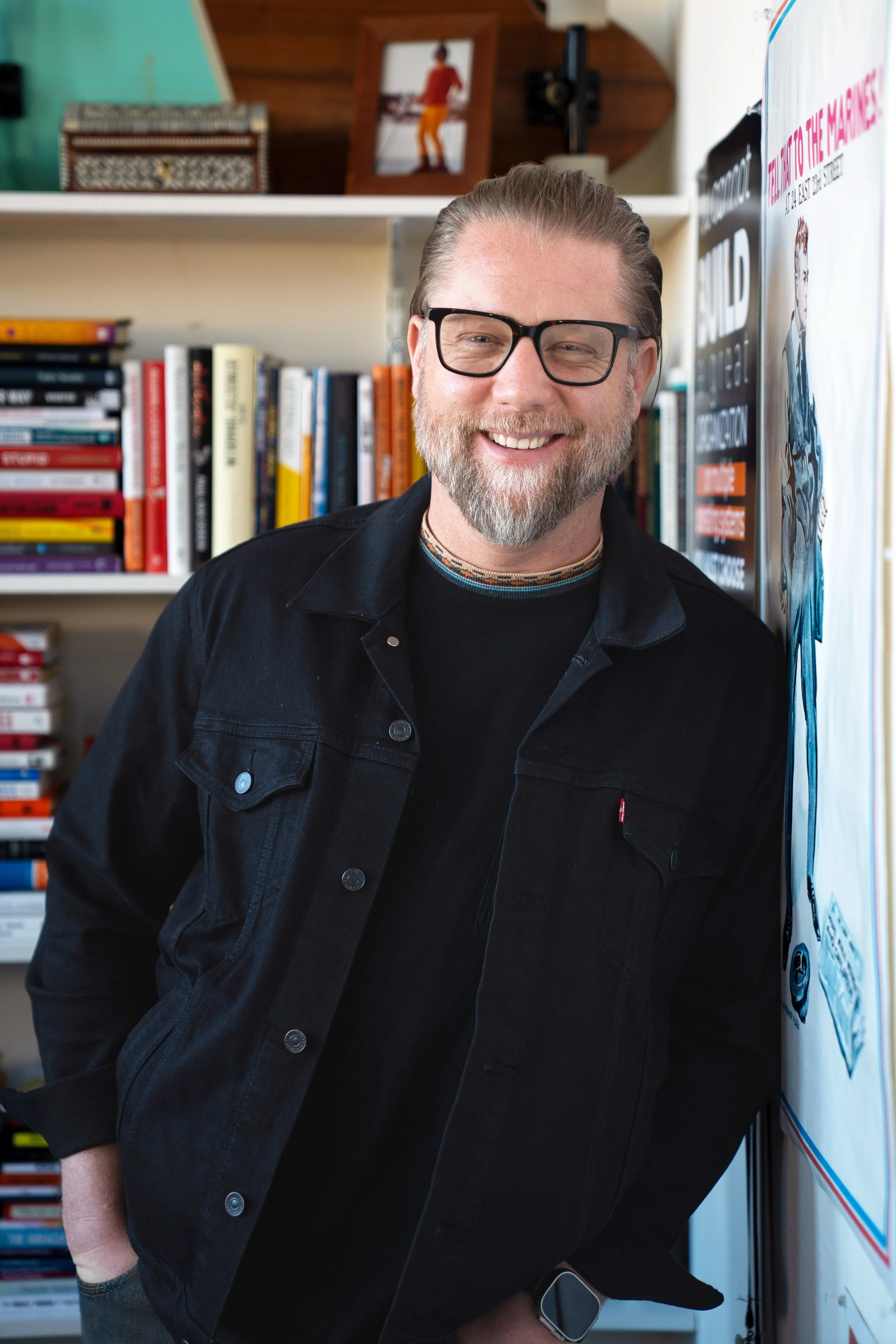 Ryan Henry smiling with glasses, a beard, and gray hair stands in front of a bookshelf filled with colorful books. He's wearing a black jacket and a smartwatch.