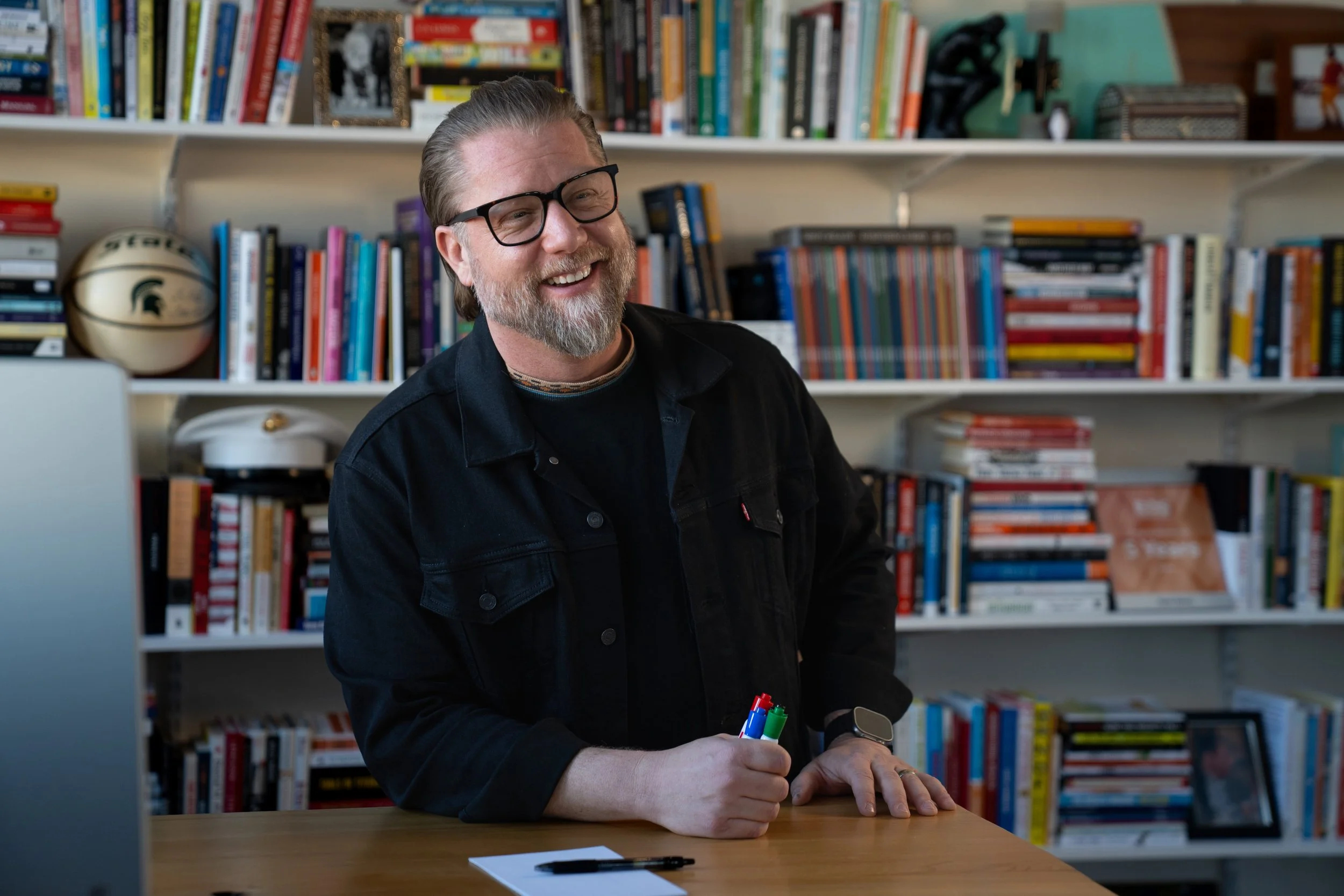 Ryan Henry with glasses and a beard, wearing a black jacket, holding colorful markers, sitting at a desk with a notebook and pen, in a room with a bookshelf filled with books and framed photos.