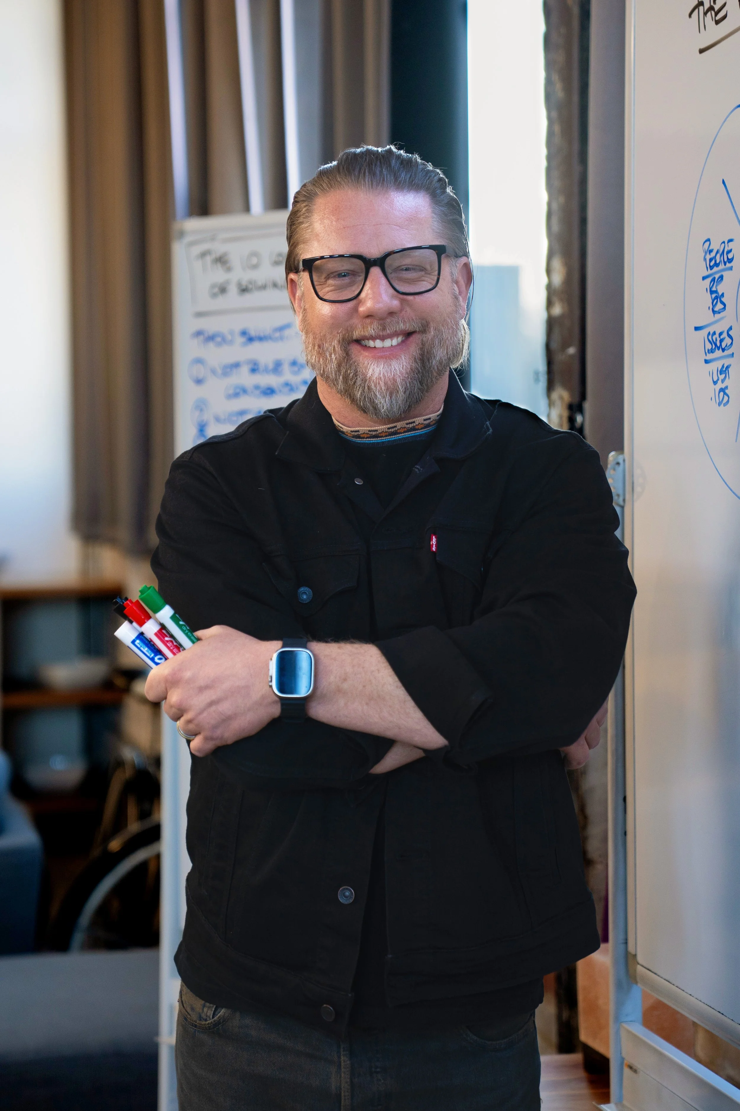 Ryan Henry smiling with glasses, a beard, and long hair tied back, standing in front of whiteboards with notes, holding colorful markers and wearing a smartwatch.