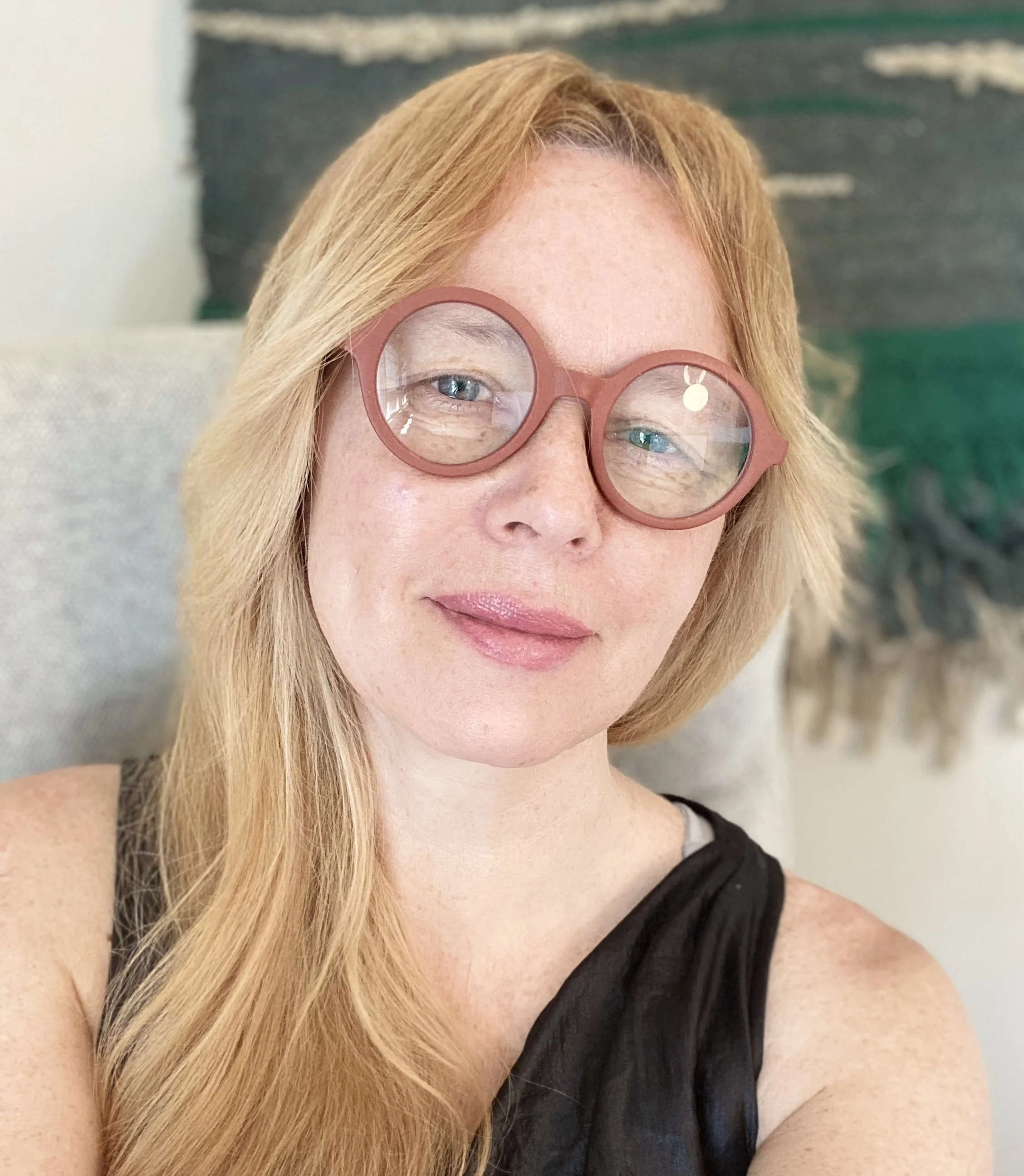 Close-up of a woman with long blonde hair wearing pink glasses and a black sleeveless top. She has light skin, blue eyes, and pink lips, and is sitting indoors with a blurred background.