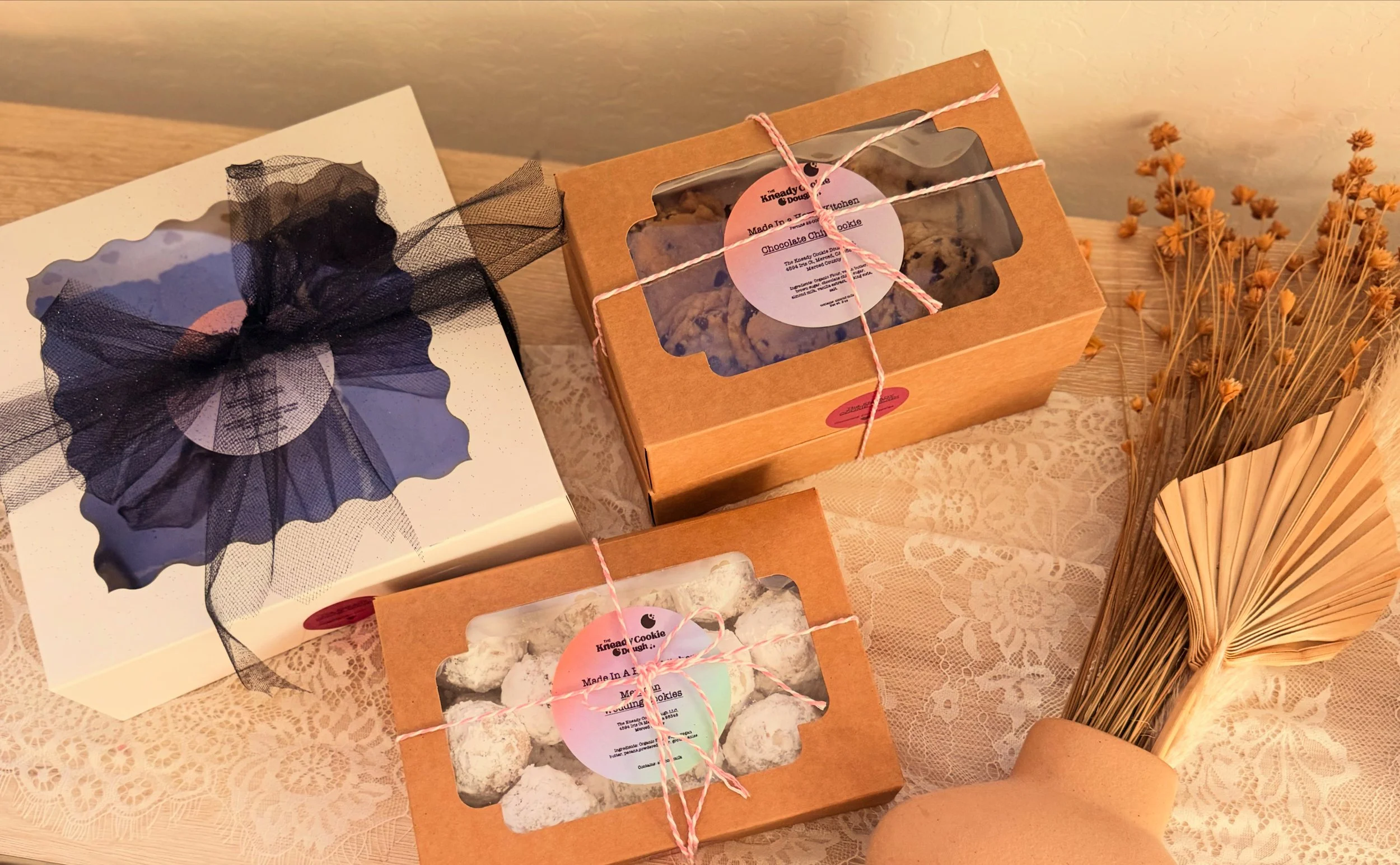 Gift boxes of assorted cookies with window panes, tied with pink and white string, a dried flower bouquet in a paper wrap, and a decorative black ribbon with a netted bow on top