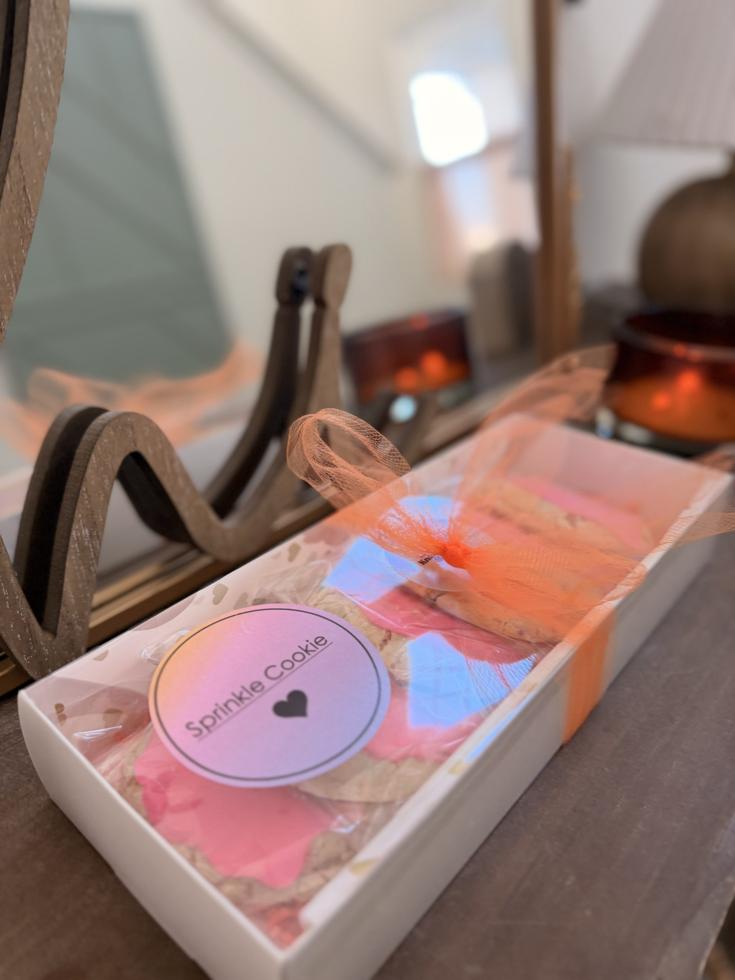 Decorative cookie packaging with a pink label that reads 'Spinkle Cookie' and a black heart, tied with an orange ribbon, placed on a wooden surface with blurred kitchen items in the background.