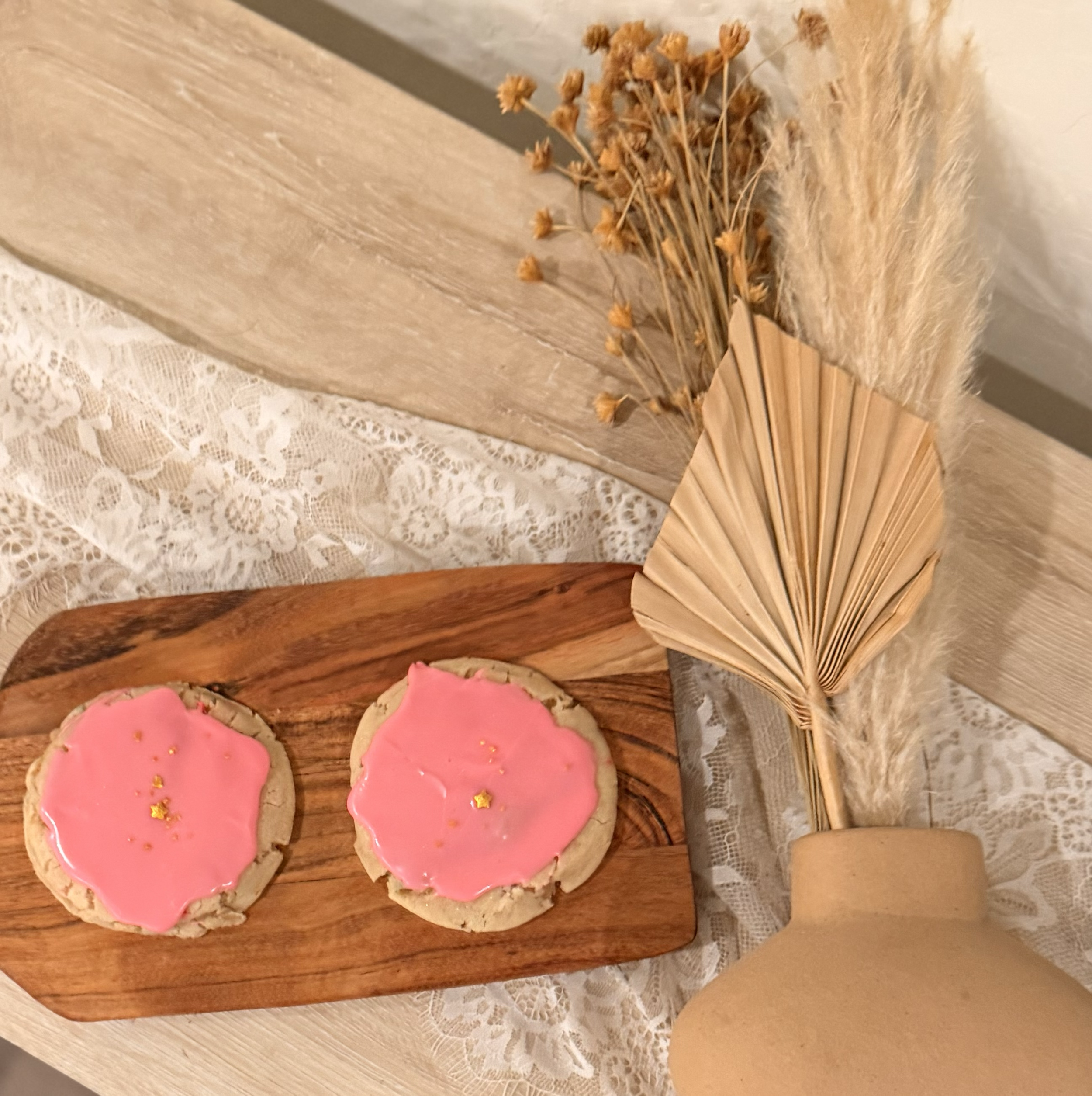 Two cookies with pink icing and gold star decorations on a wooden tray, next to a dried floral arrangement and a beige vase, all on a lace tablecloth.