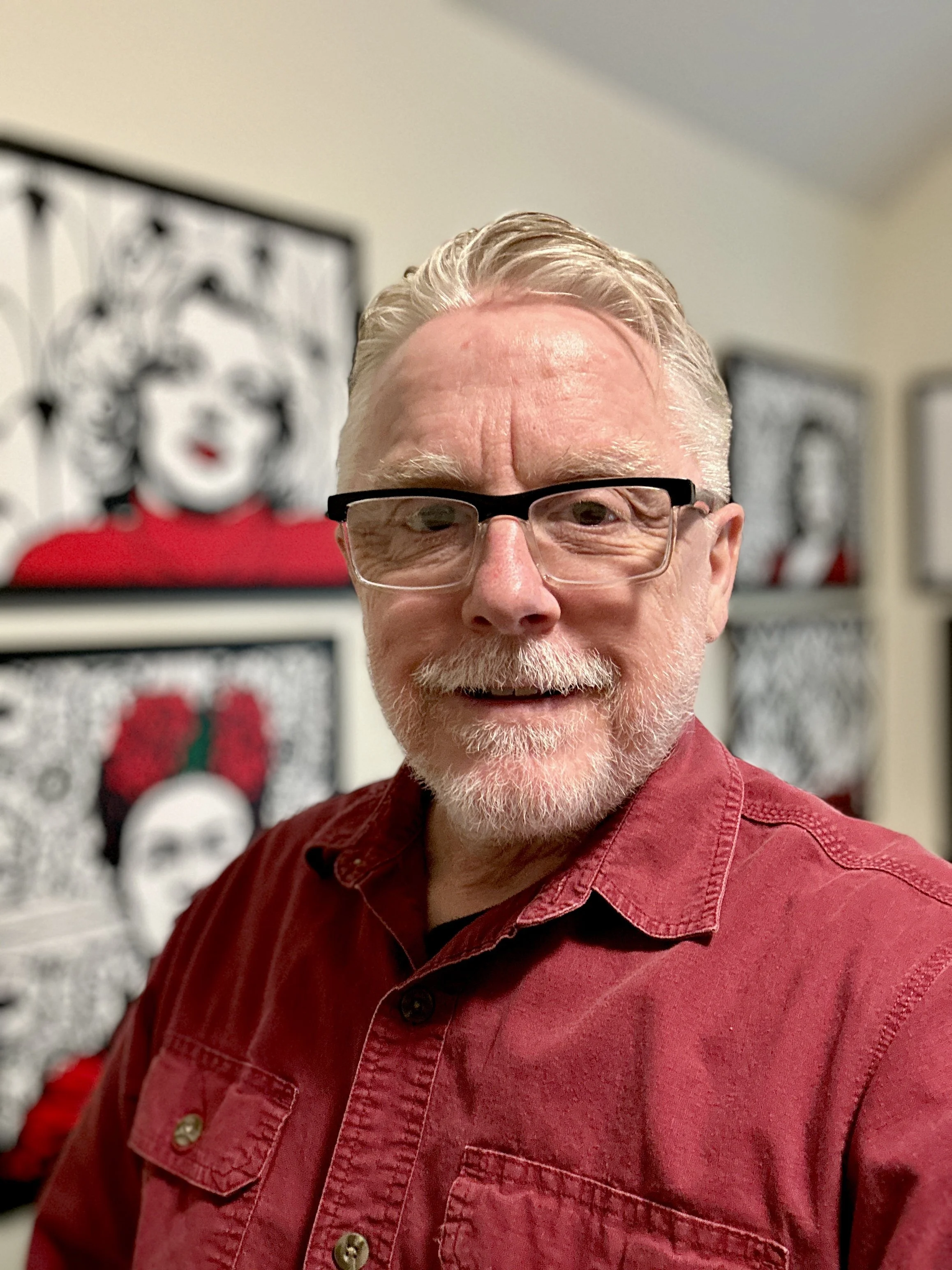 Close-up image of an older man with glasses and gray hair, smiling and wearing a red shirt, with art featuring a portrait of a woman in the background.
