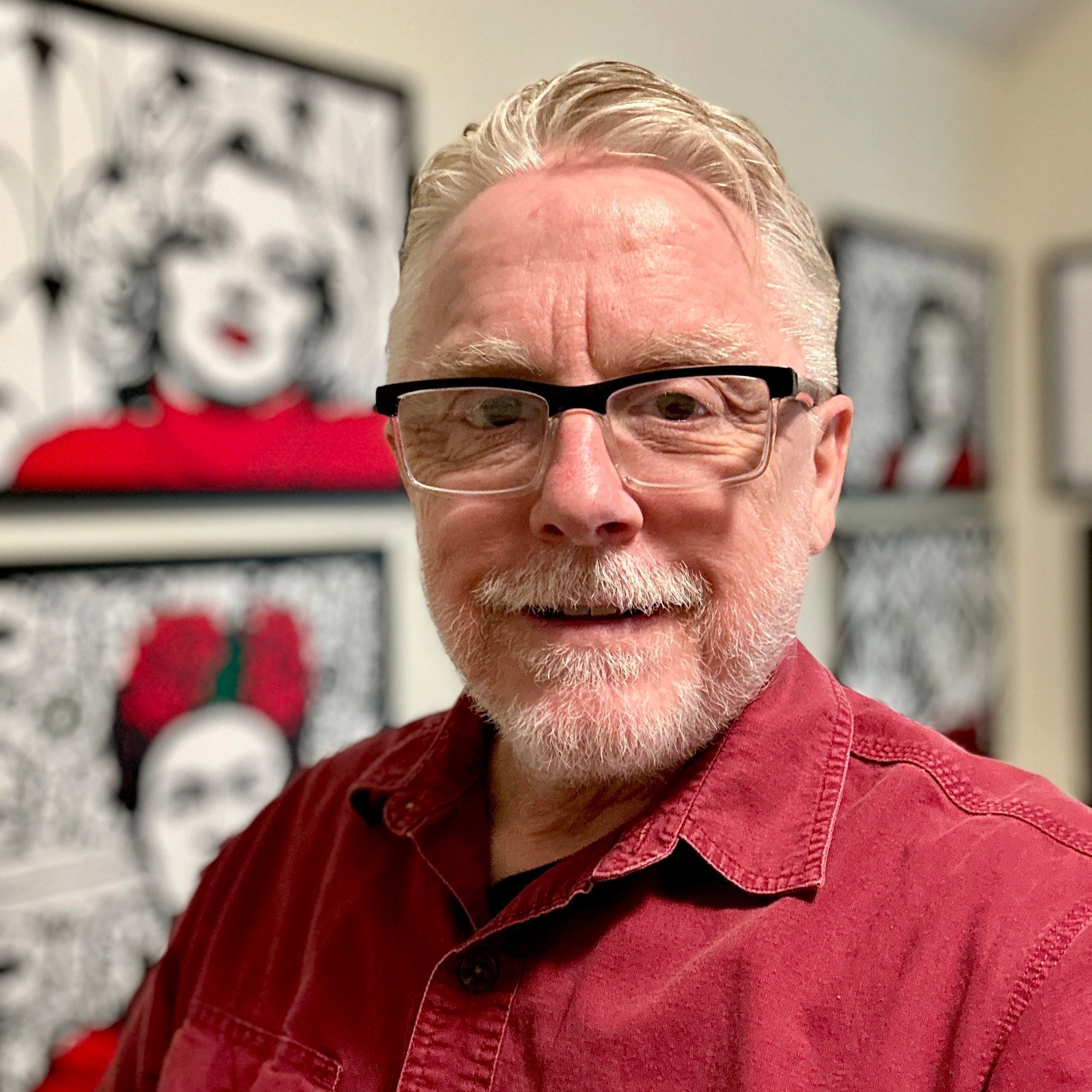 Close-up of a man's face wearing glasses, with white hair, beard, and mustache, smiling slightly, wearing a red shirt, with blurred holiday decorations in the background.