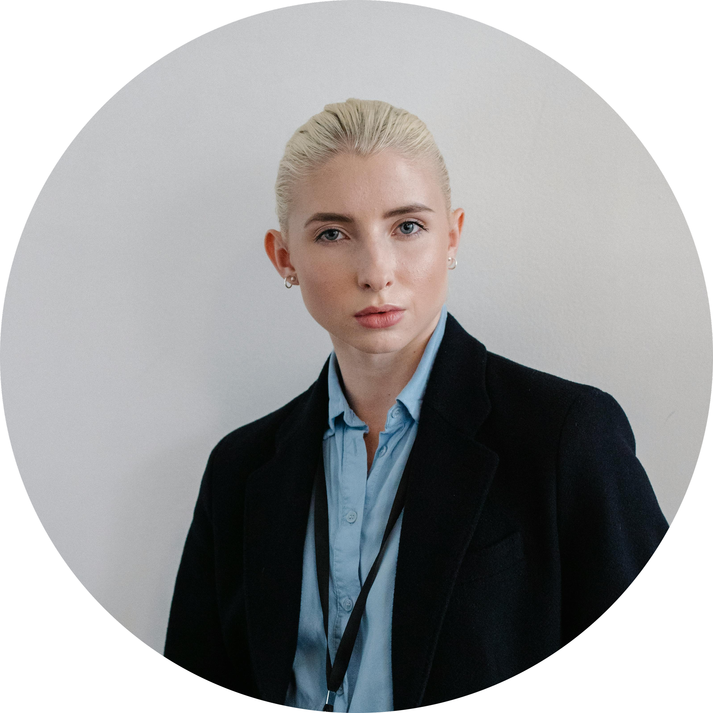 Portrait of a young woman with blond hair tied back, wearing a black blazer and light blue collared shirt, looking directly at the camera against a plain white background.