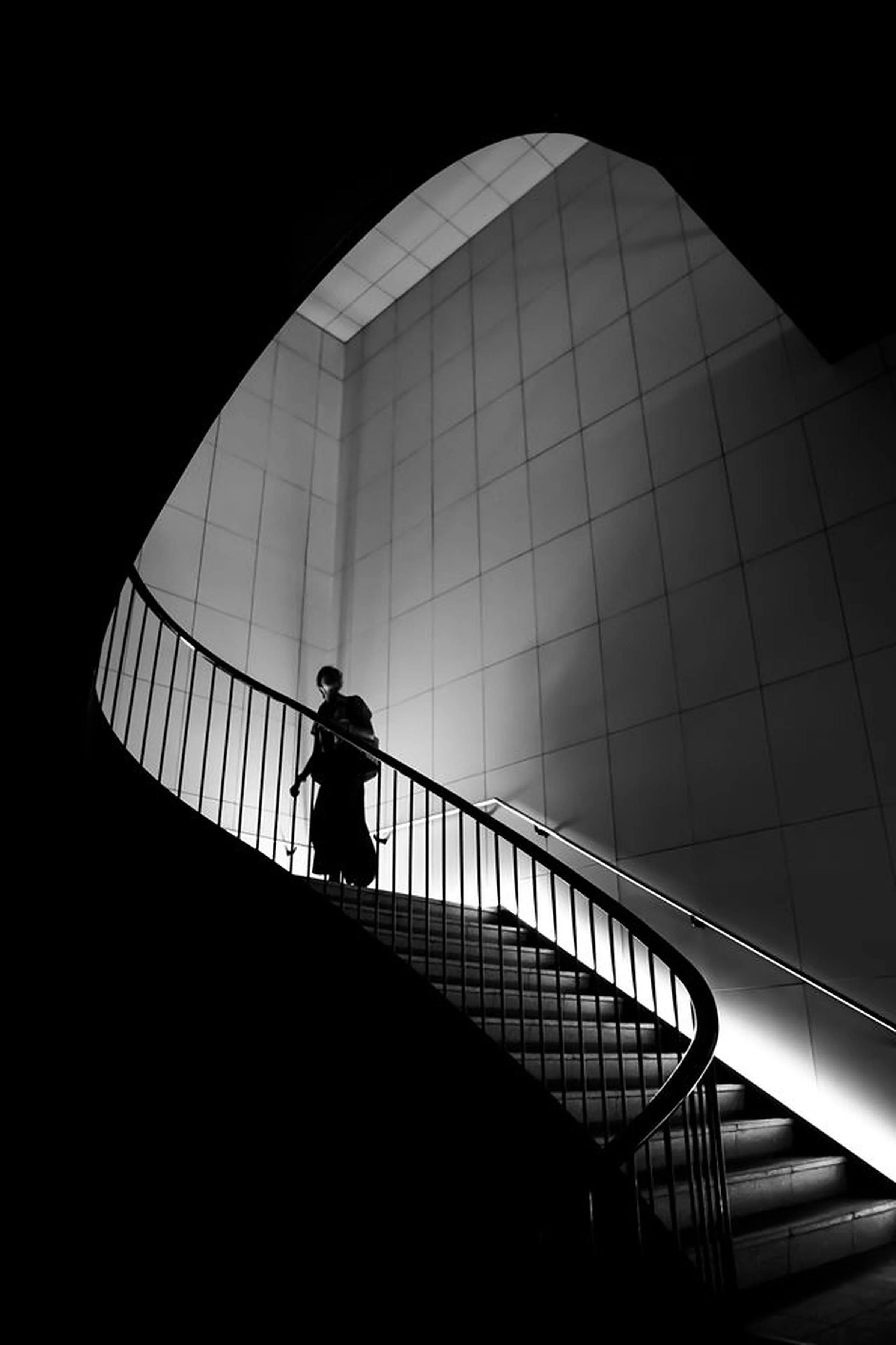 A black-and-white photo of a person walking up a curved staircase with modern design, railing, and textured wall background.