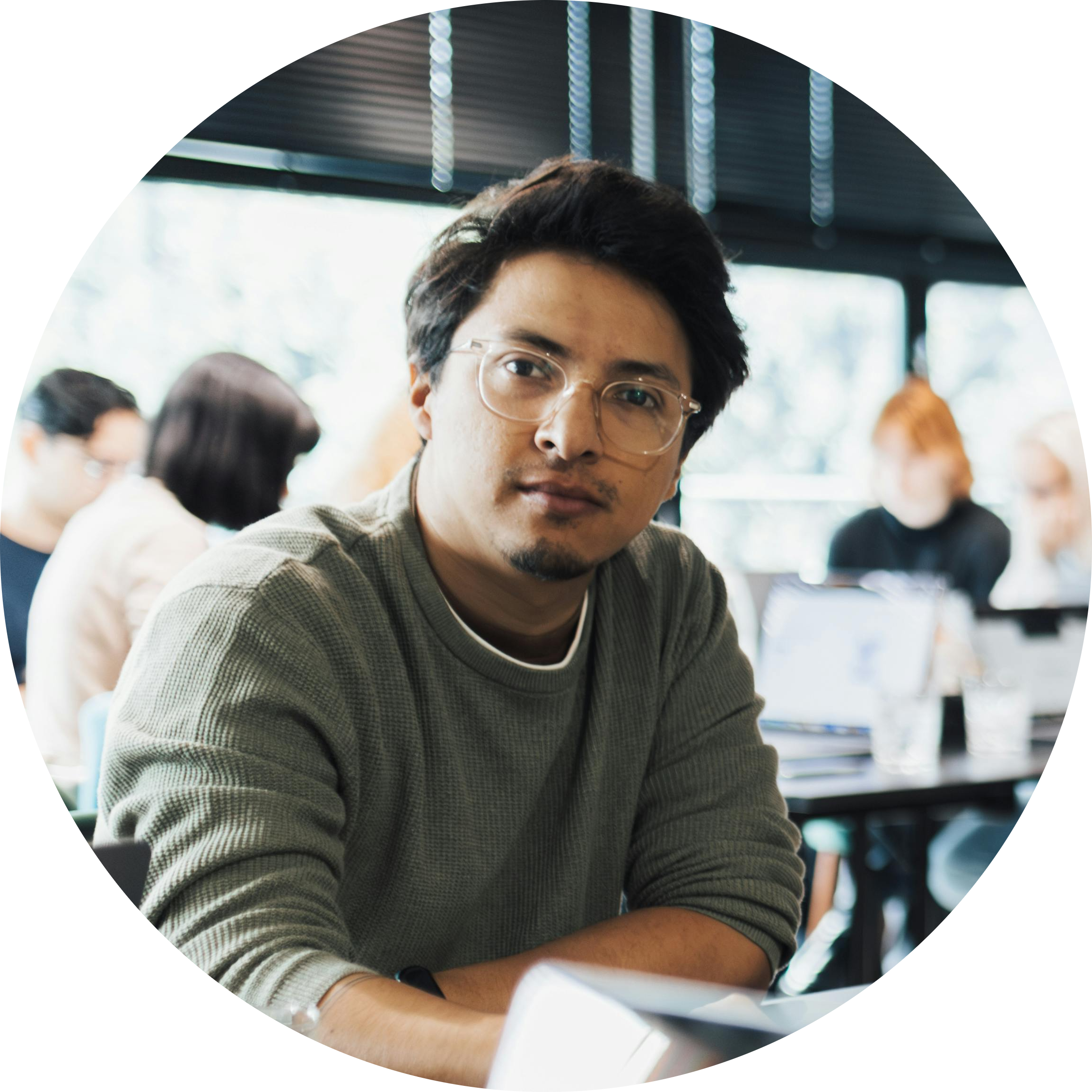 A young man with dark hair, glasses, and a goatee looking directly at the camera in a busy indoor setting with others working in the background.